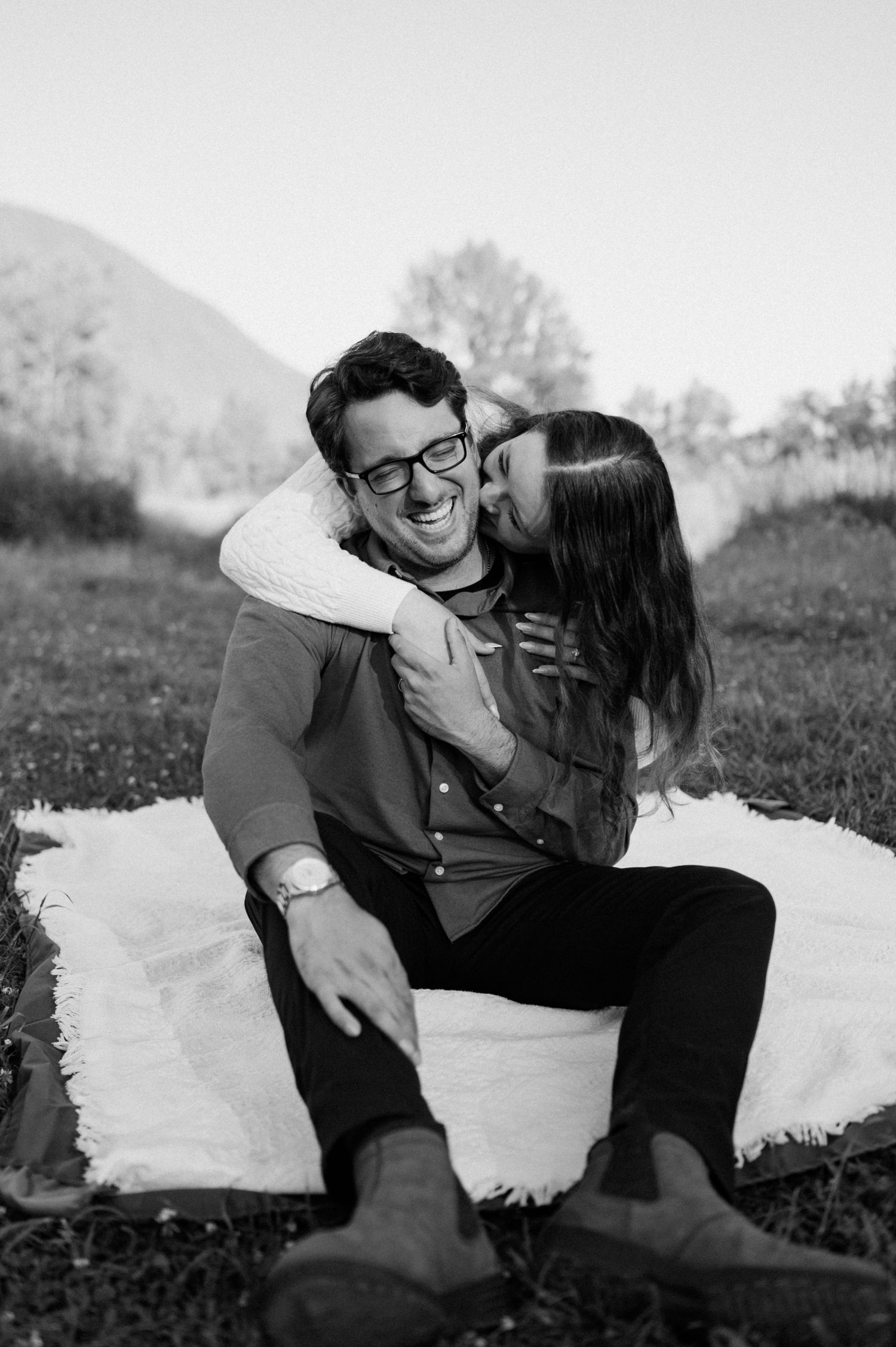 A couple sharing an affectionate moment on a white blanket with mountains in the background.