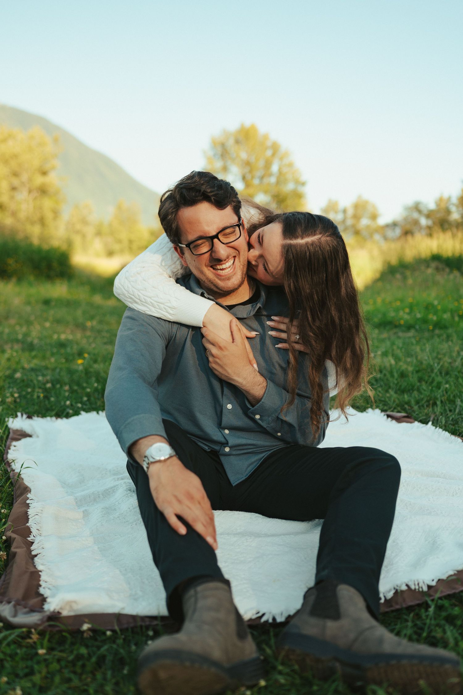 A tender moment shared between a couple sitting on a white blanket in a mountain meadow.