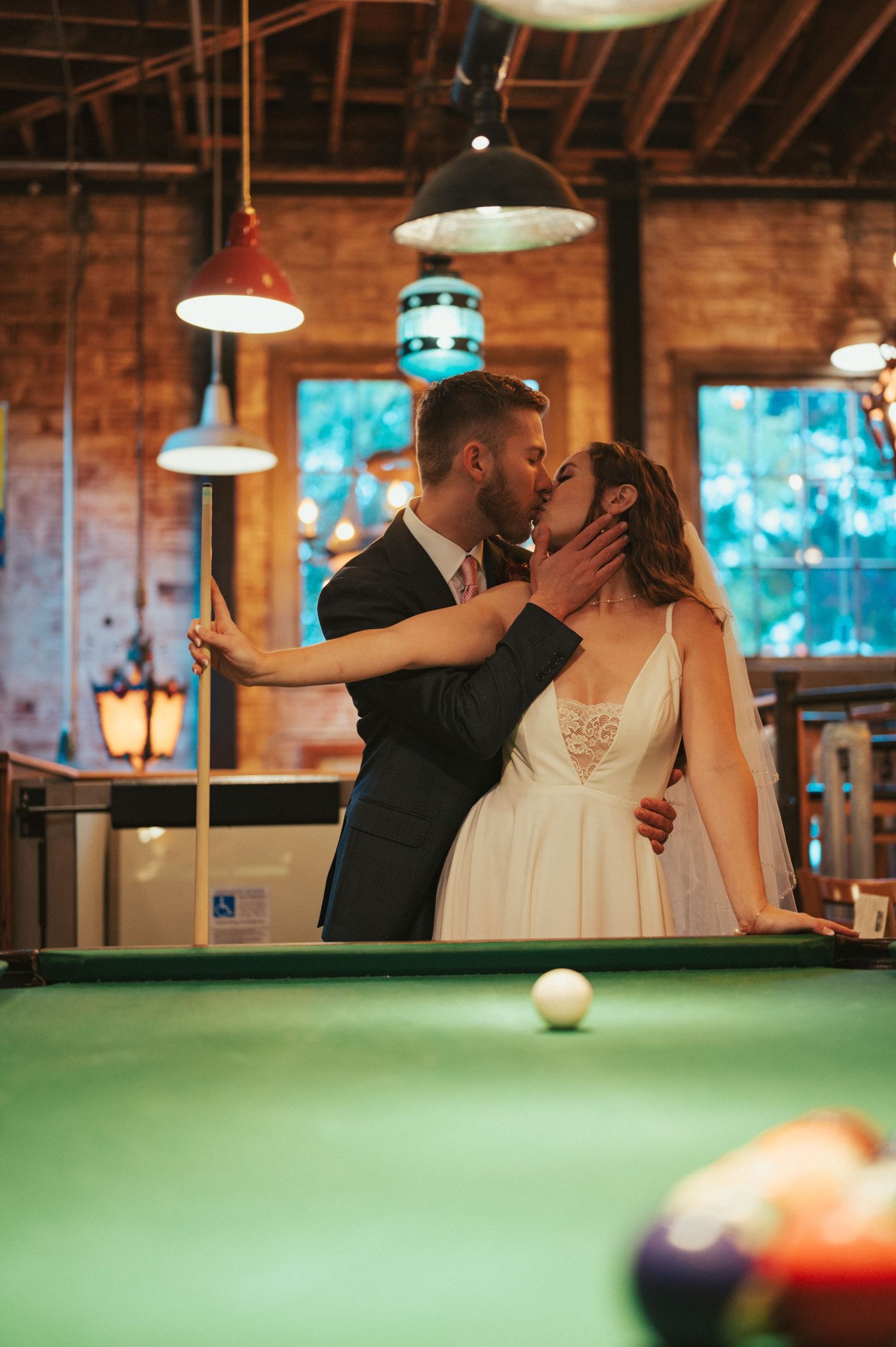 A bride and groom share a romantic moment by a pool table in a rustic venue with hanging pendant lights and exposed brick.