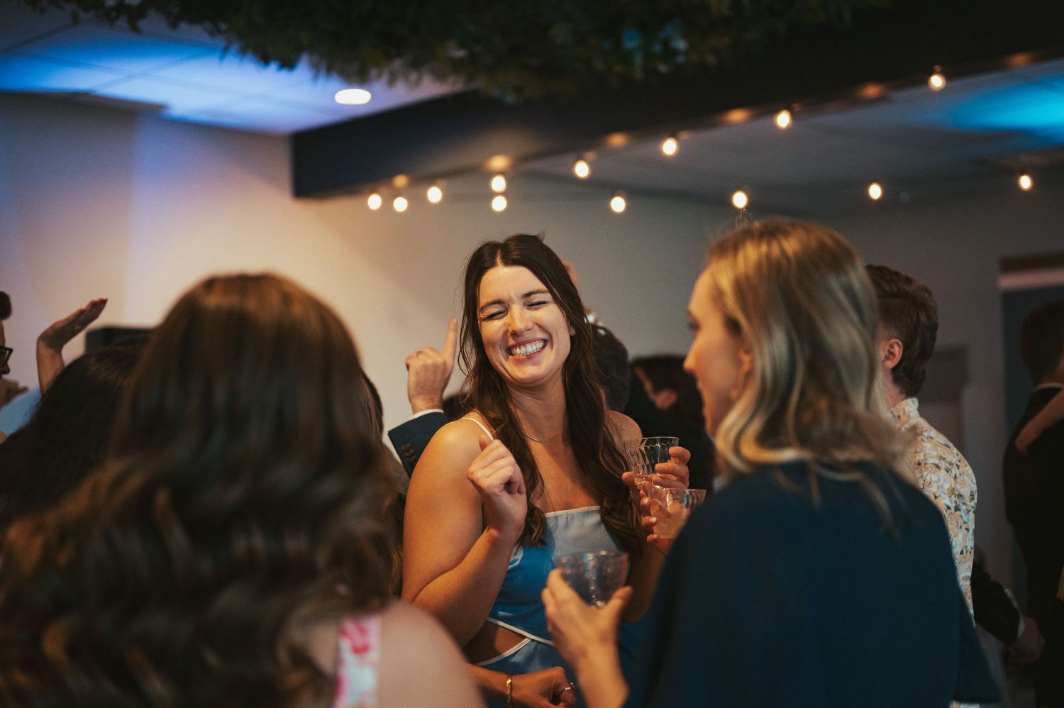 String lights illuminate a dark reception venue as guests dance and celebrate.