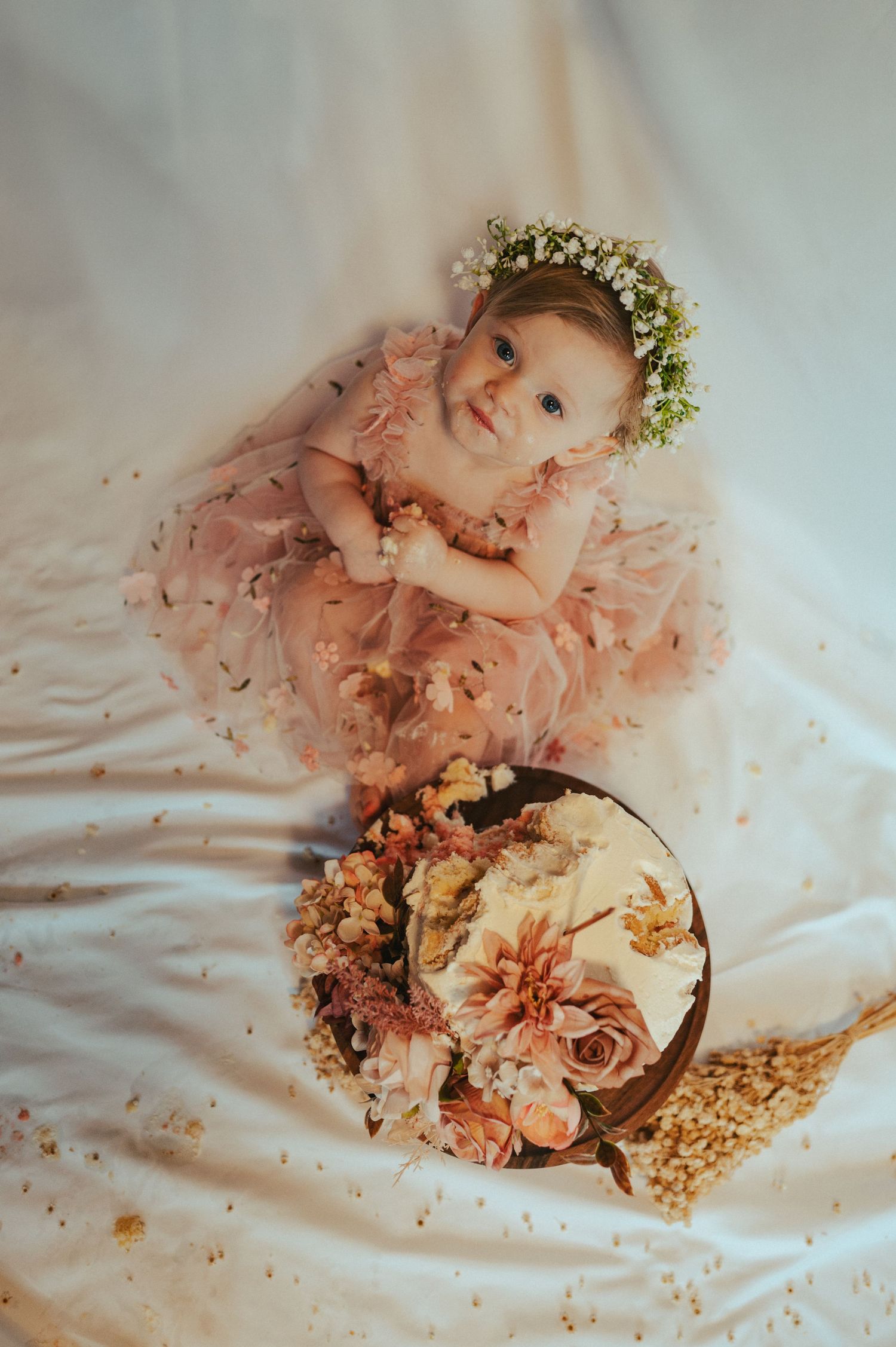 A baby in a flower crown and pink dress poses with floral decorations during a photoshoot.