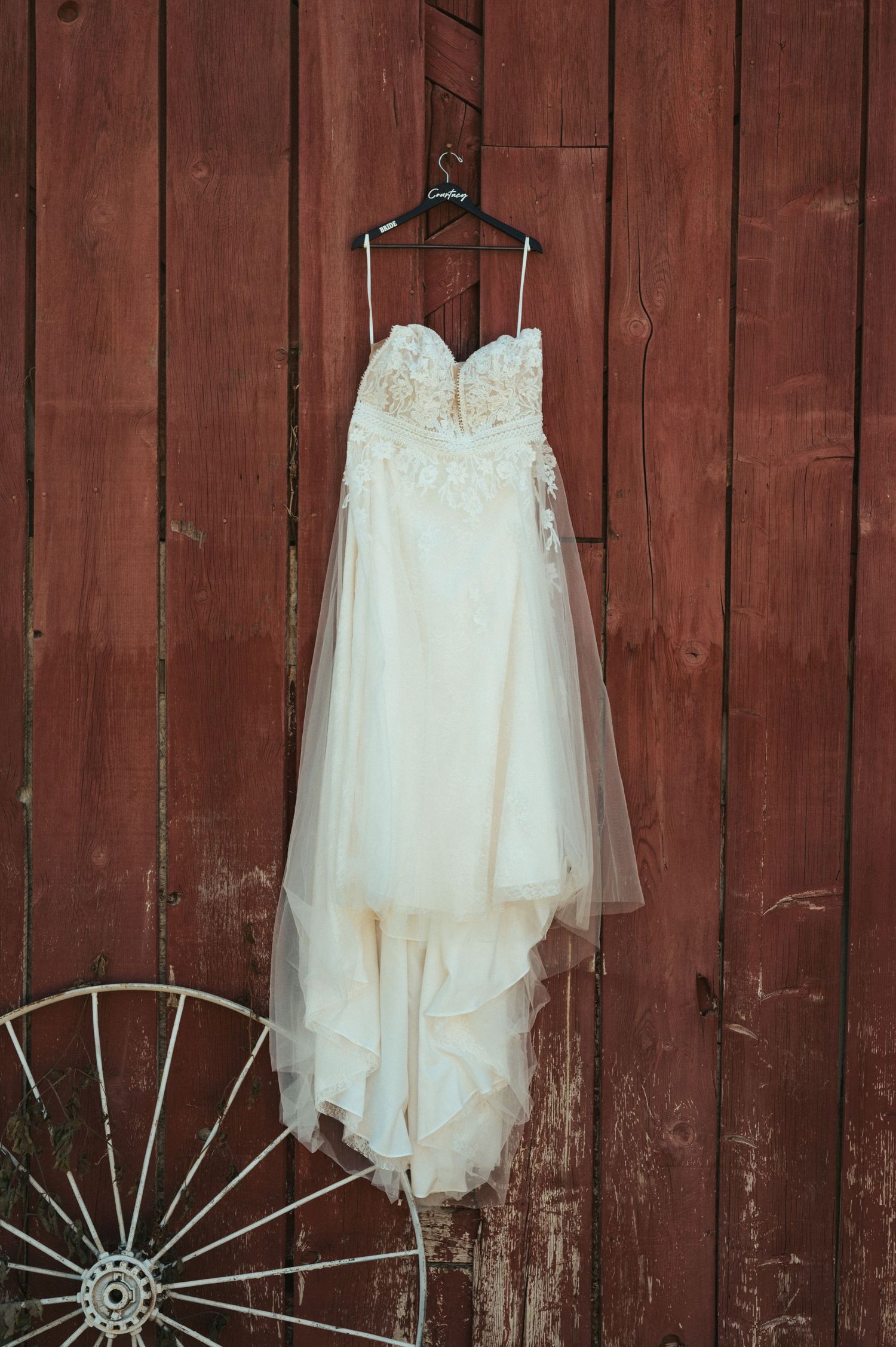 White lace wedding dress hangs on rustic red barn wall beside vintage wagon wheel.