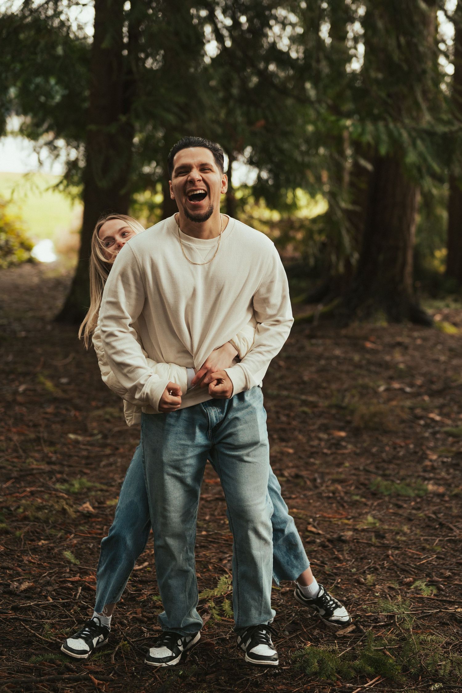 Couple in matching white sweaters and jeans share playful moments in a wooded setting.
