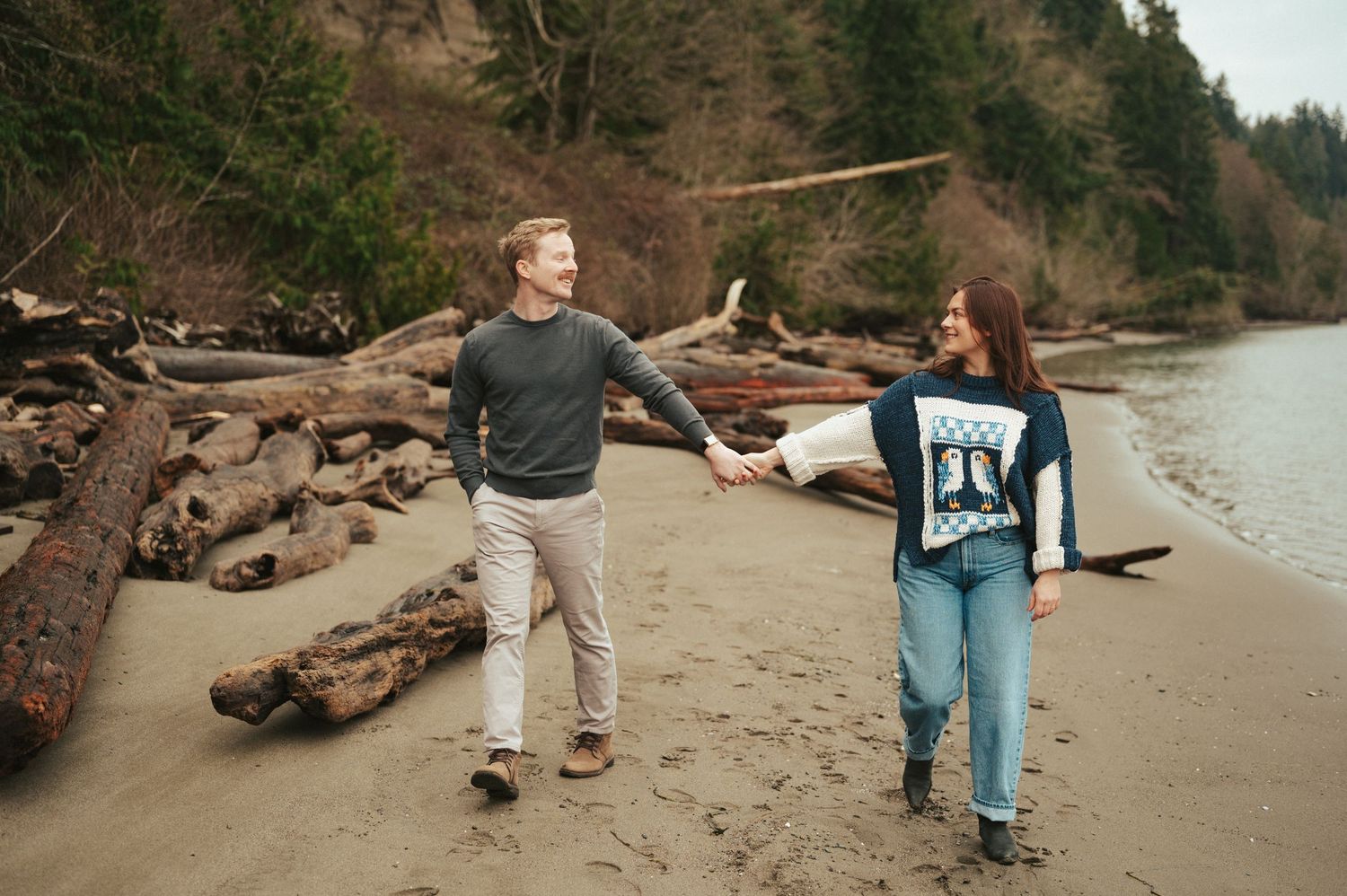 A playful couple walking on a beach with driftwood logs and evergreen trees.