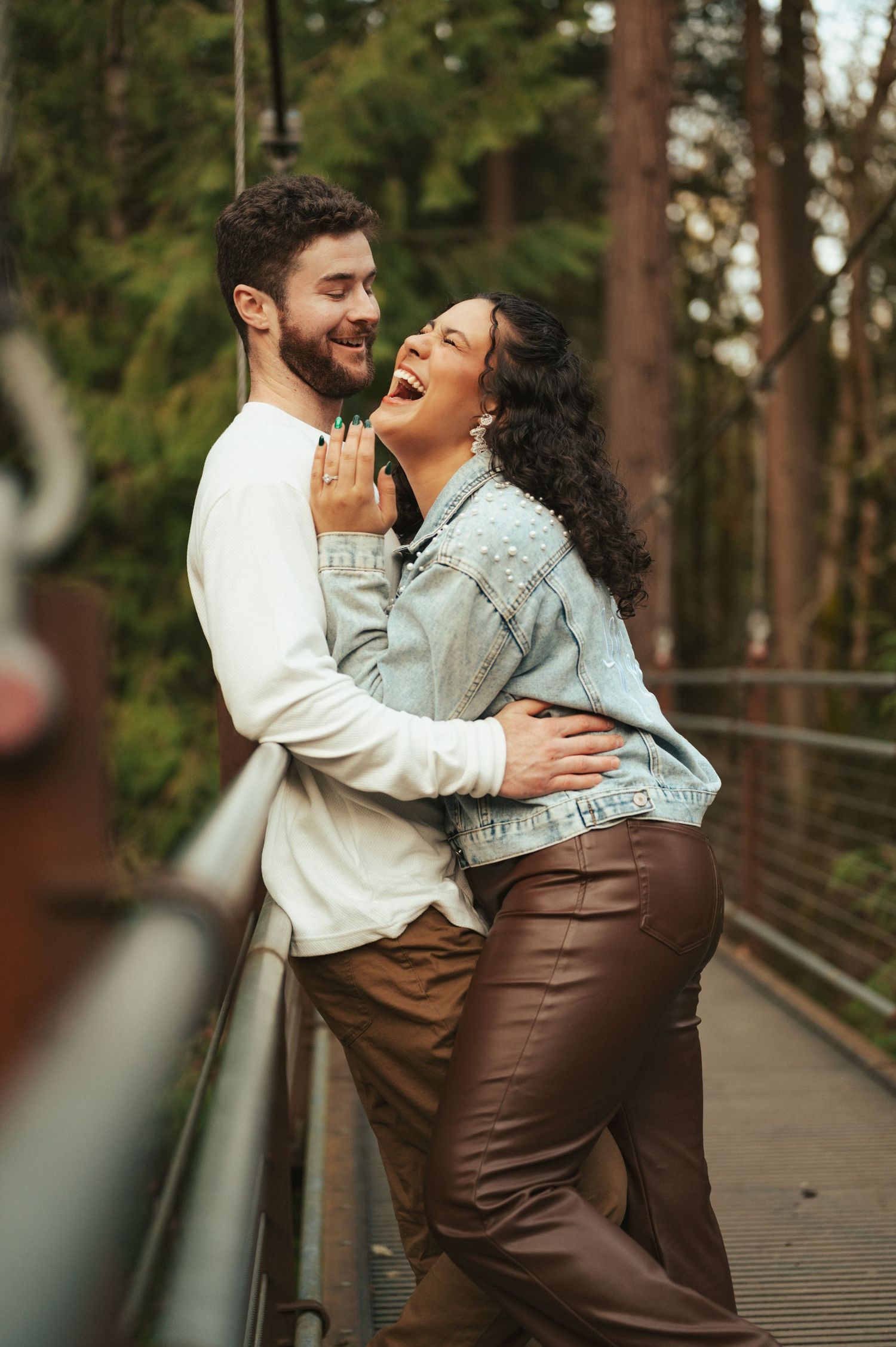 Joyful couple laughing and embracing on a wooden bridge surrounded by trees.