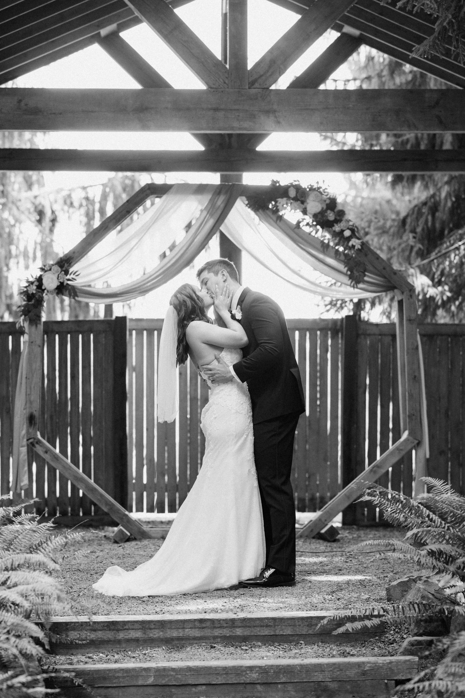 Black and white photo of romantic wedding kiss under wooden arch with draped fabric.