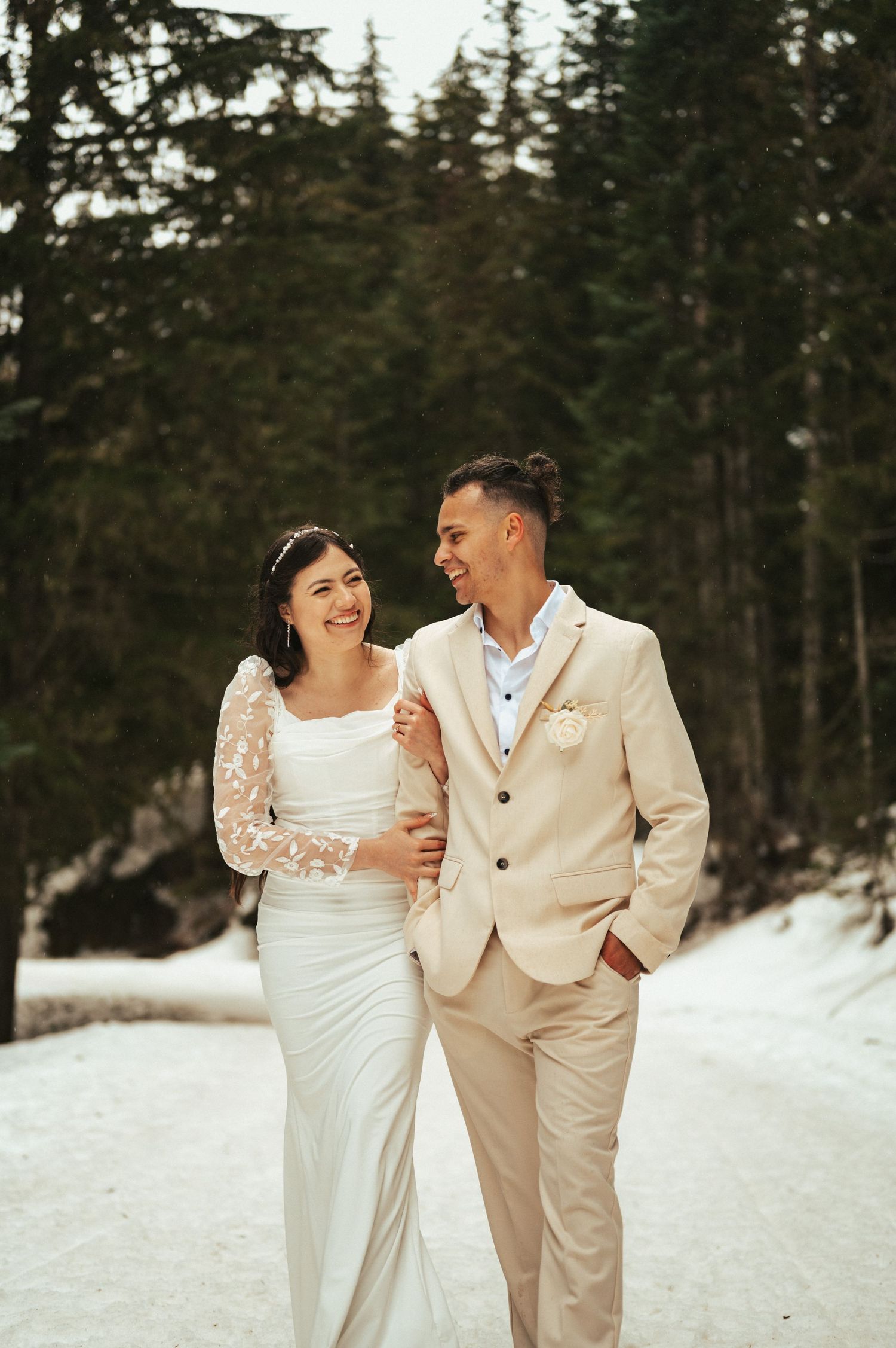 Happy couple in winter wedding attire walking through snowy forest setting.