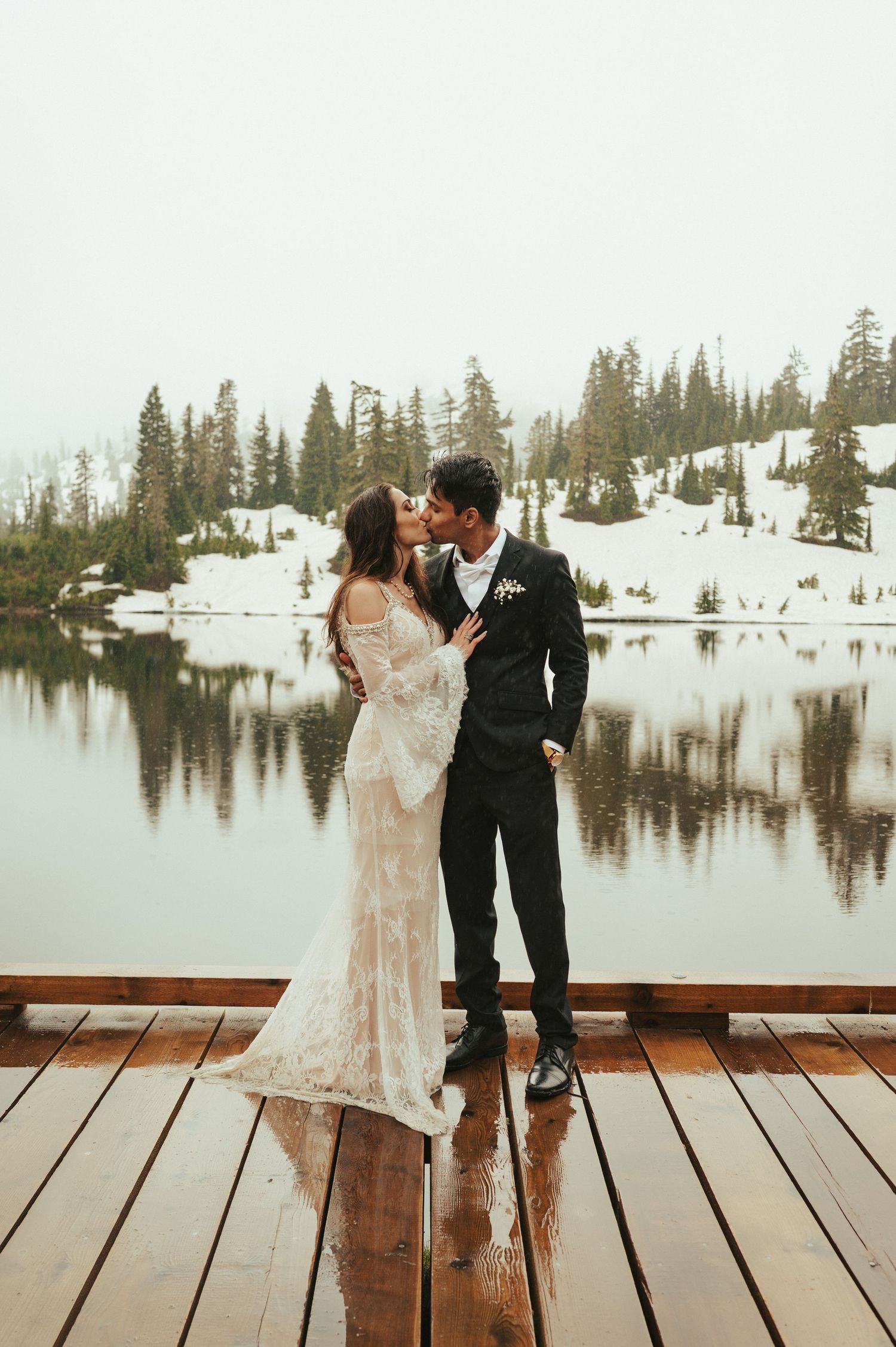 Newlyweds share an intimate moment on a dock with a snowy mountain lake backdrop.