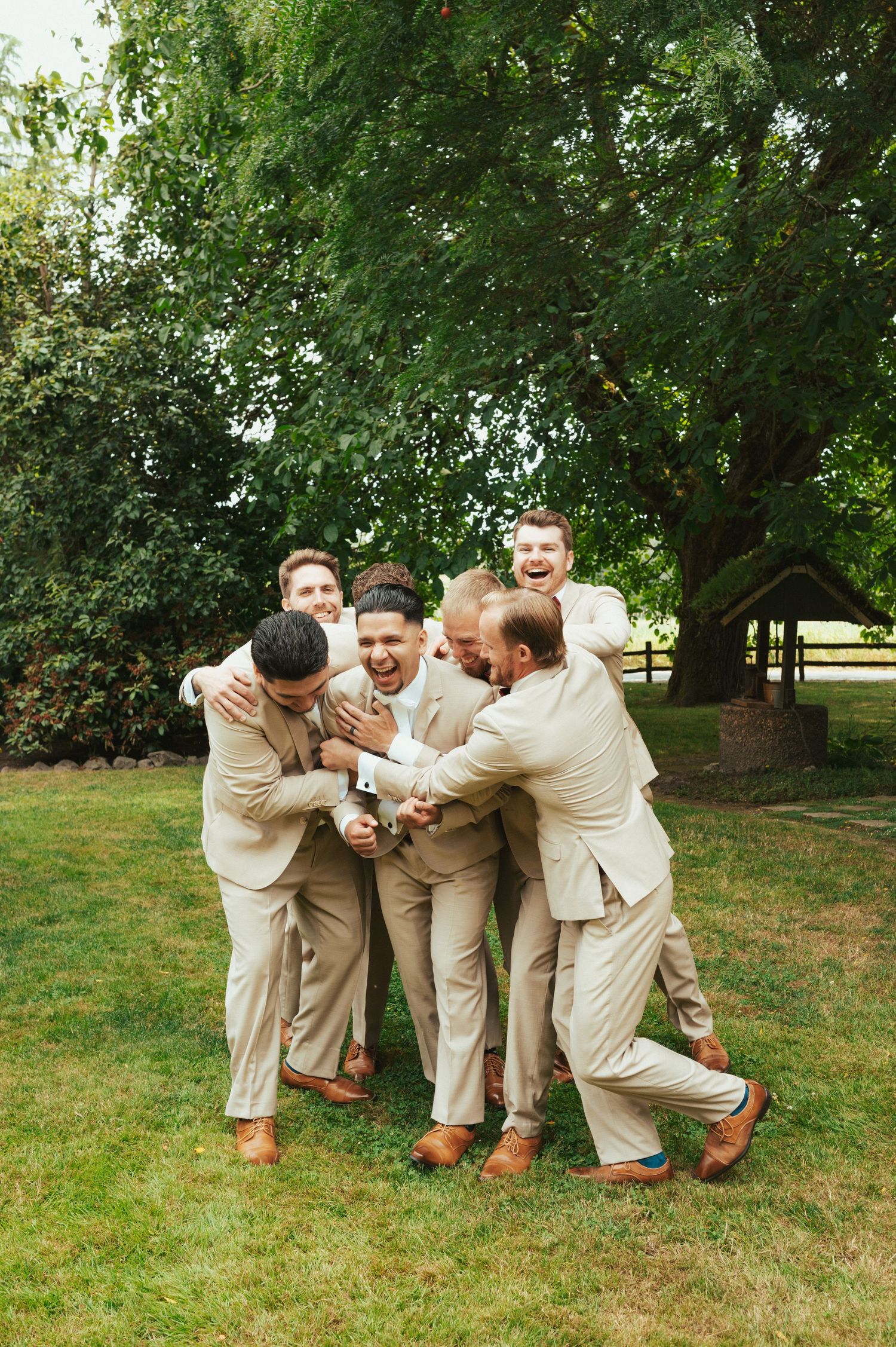 Groomsmen in beige suits playfully huddle together on a sunny lawn during wedding celebrations.