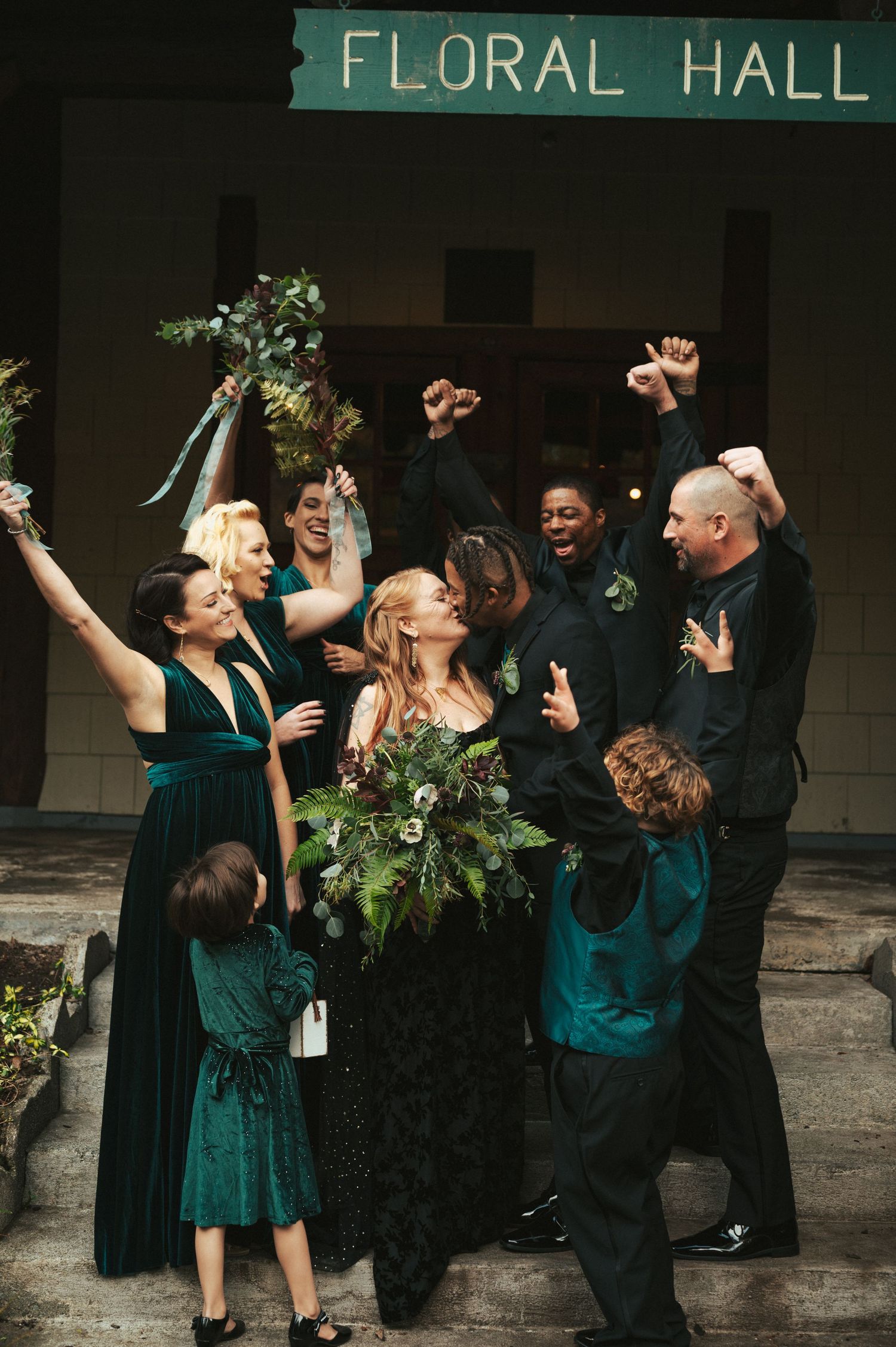 A joyful wedding party celebrates on the steps of Floral Hall with raised hands and bouquets.
