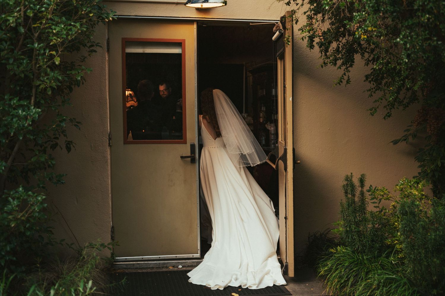 Bride in flowing white gown enters doorway with dramatic lighting.