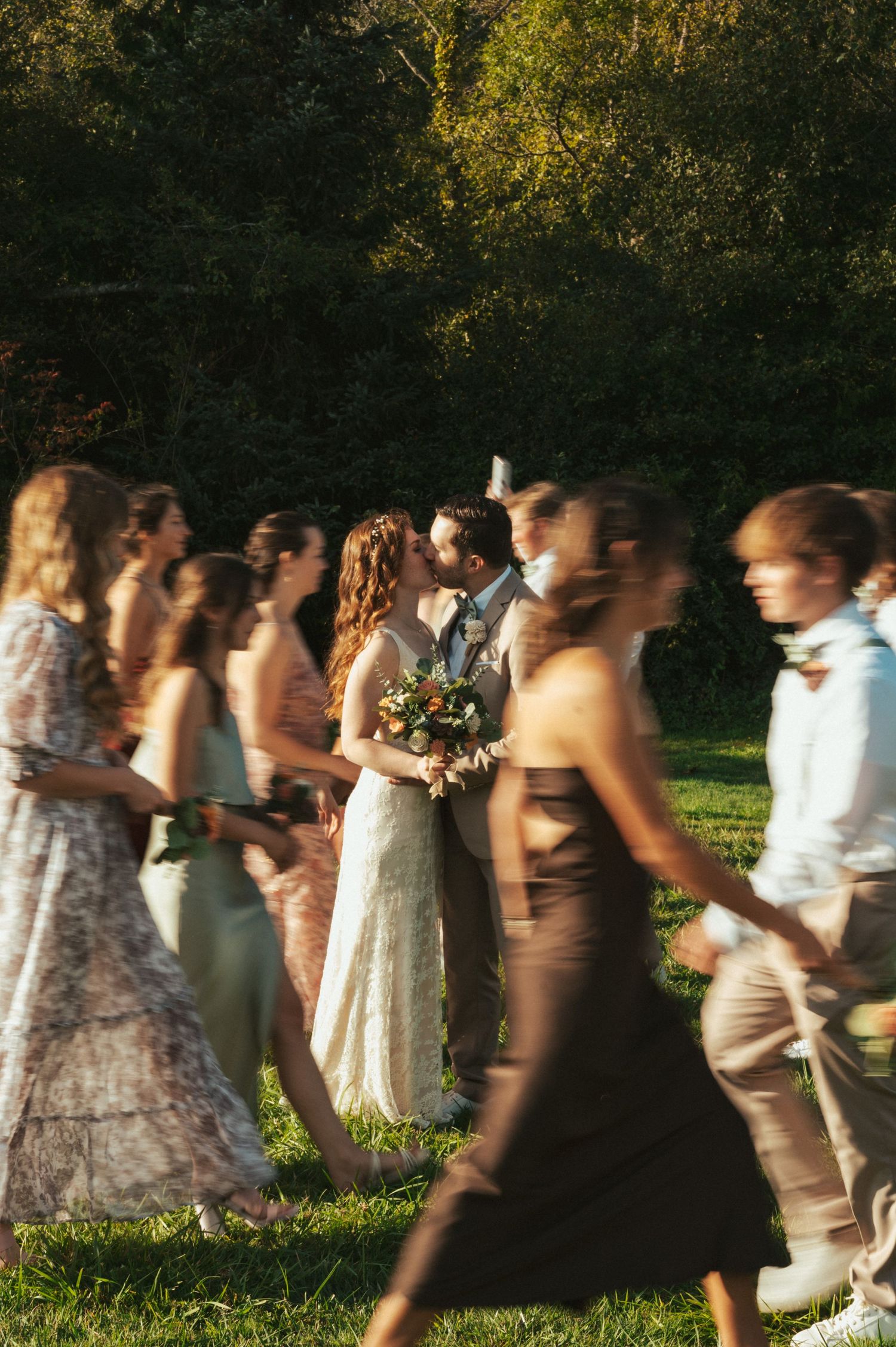 A wedding party walks across a sunlit lawn with bridesmaids in flowing dresses.