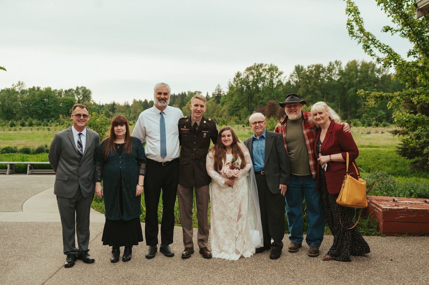 A group of wedding attendees pose outdoors on a sunny day with trees and greenery visible in the background.