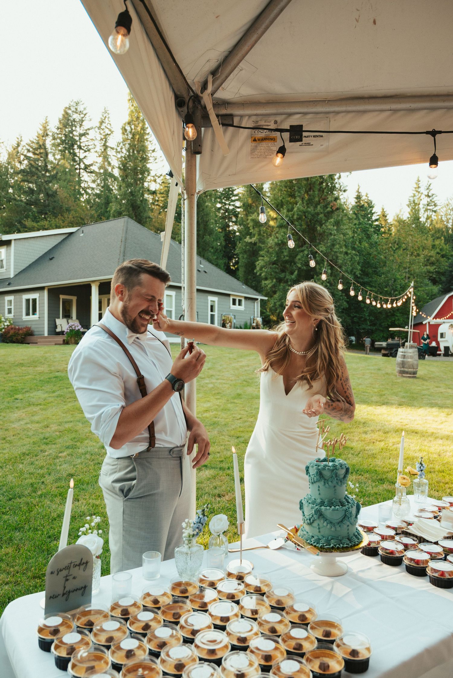 Couple sharing cake at outdoor wedding reception with dessert table.