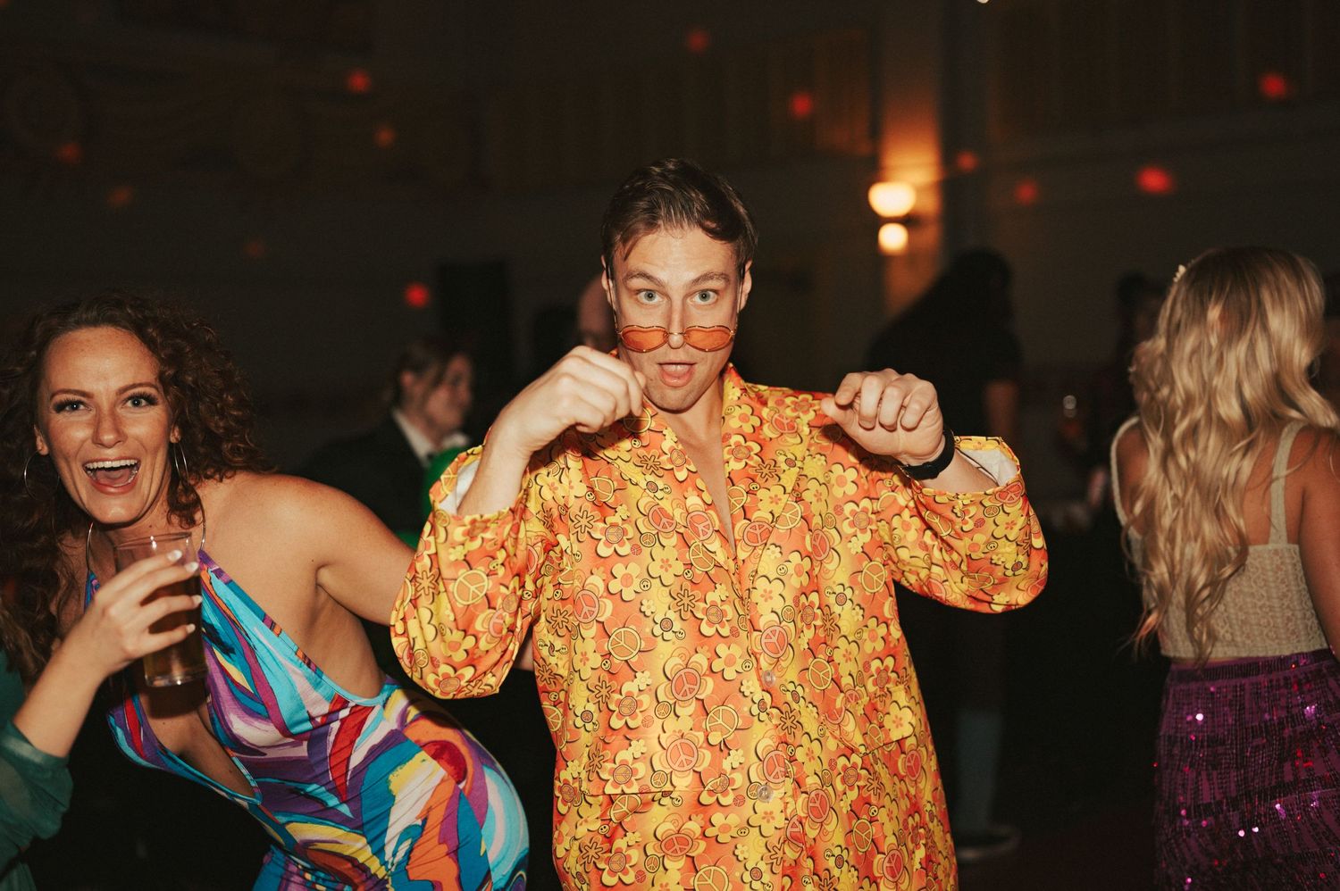 People dancing and having fun at a party with someone wearing a bright orange patterned shirt in focus.