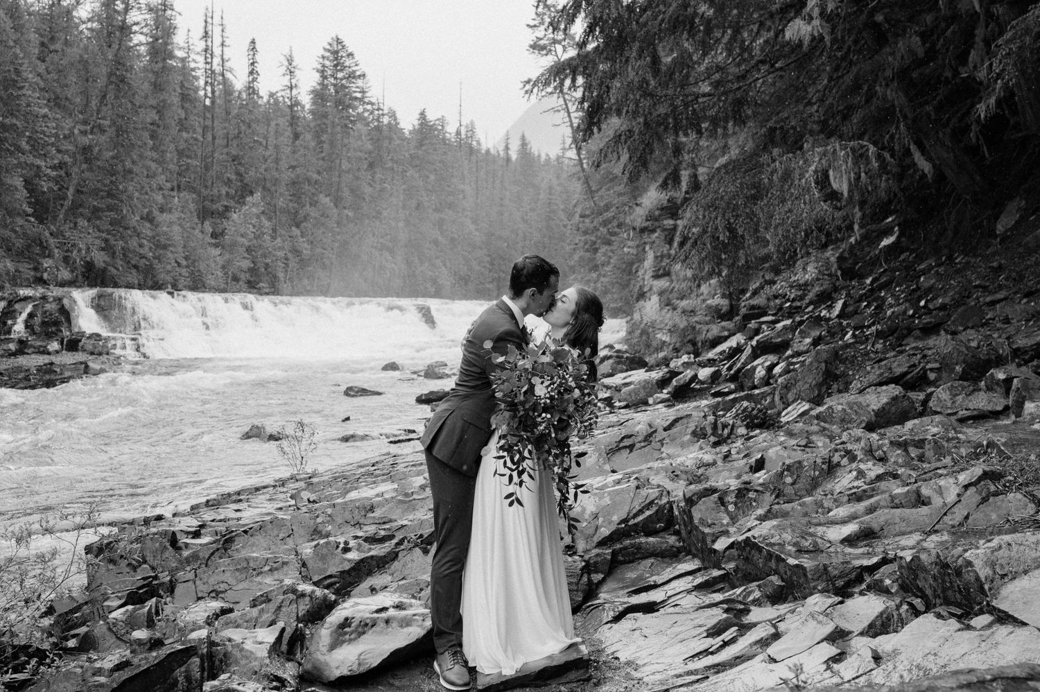 A couple embraces near a scenic waterfall on rocky terrain in a black and white nature setting.