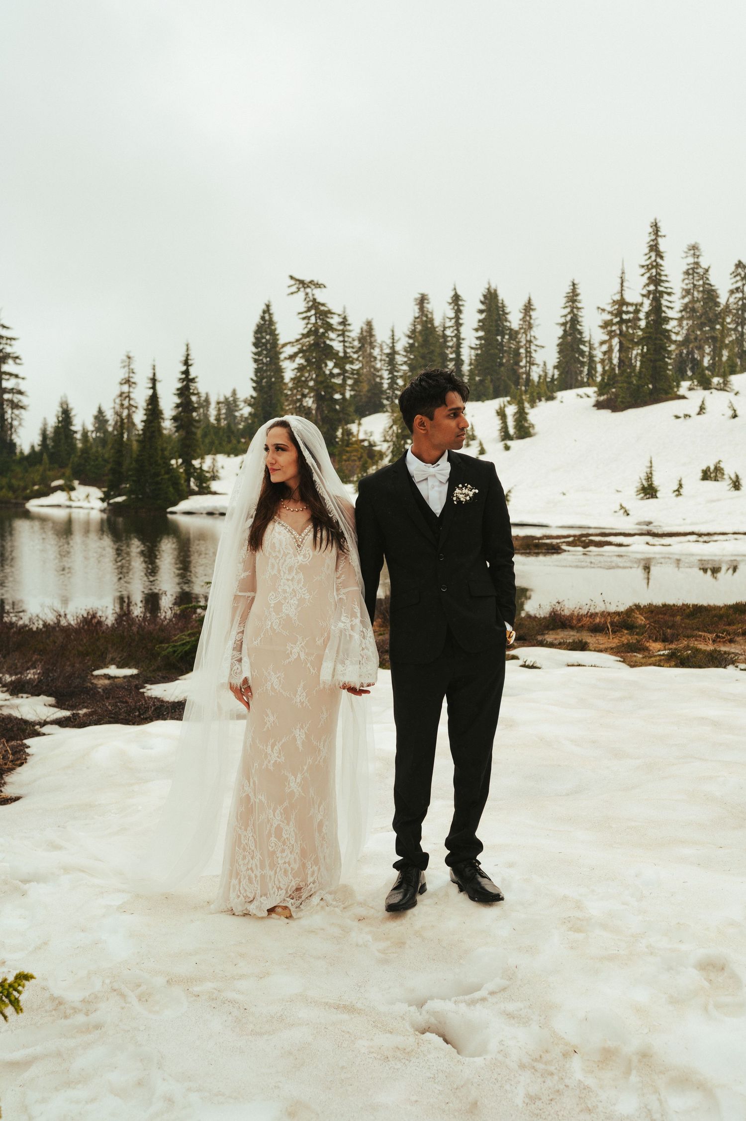 Wedding couple poses in front of a snow-covered mountain landscape with evergreen trees.
