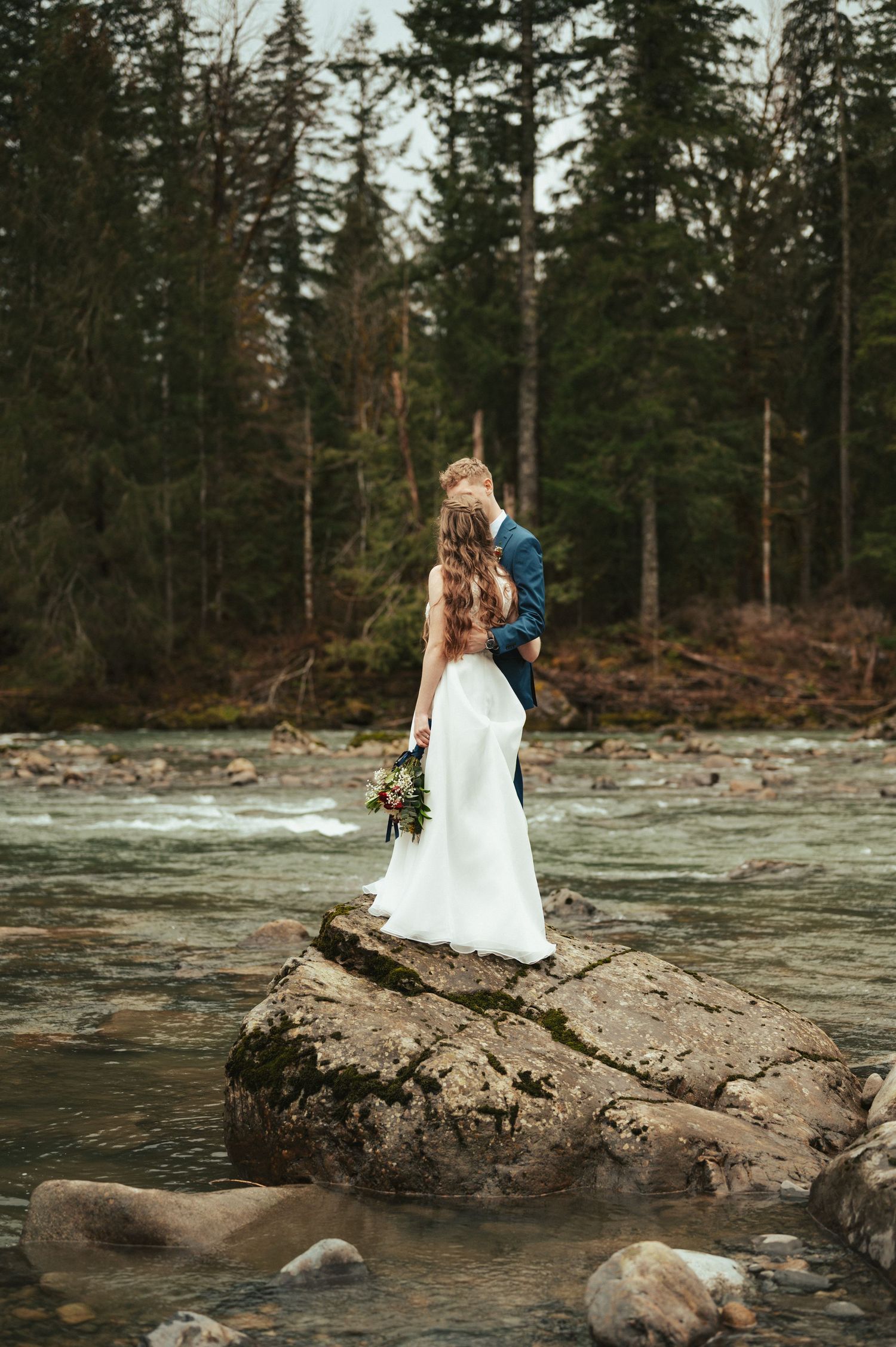 Bride in white dress stands on rocky outcrop by frozen stream in evergreen forest.