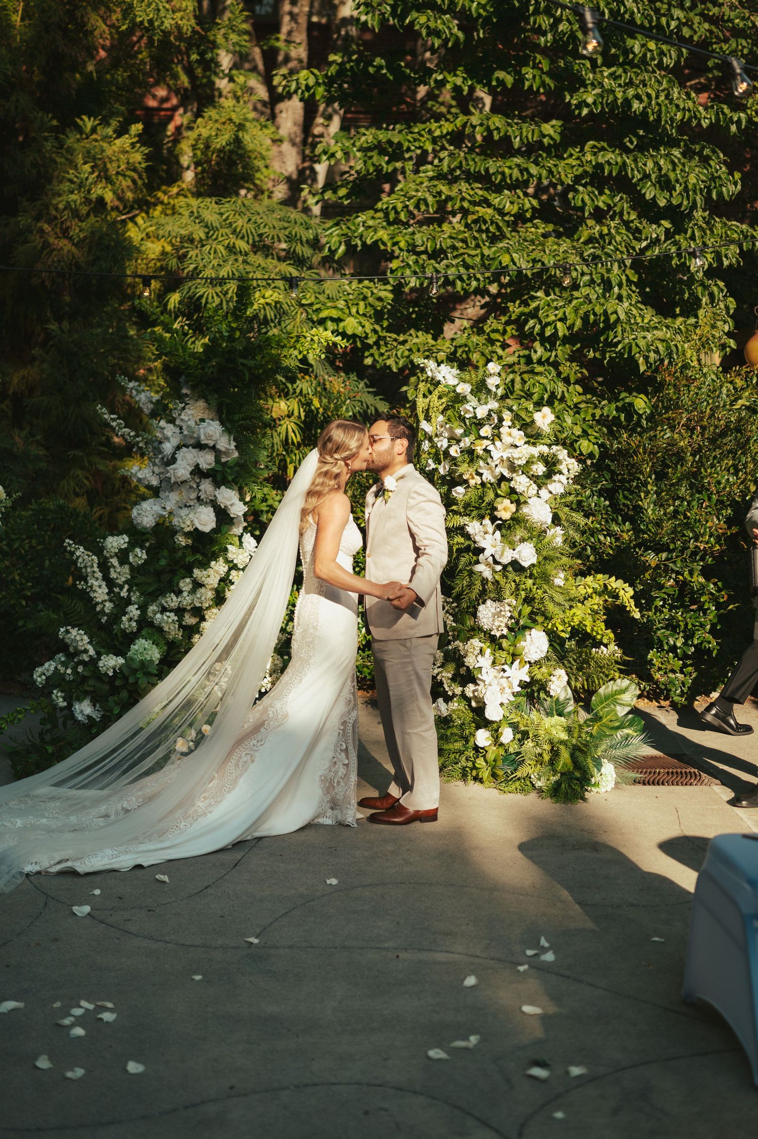 Romantic wedding couple shares intimate moment in lush garden setting with white floral arrangements and flowing veil.