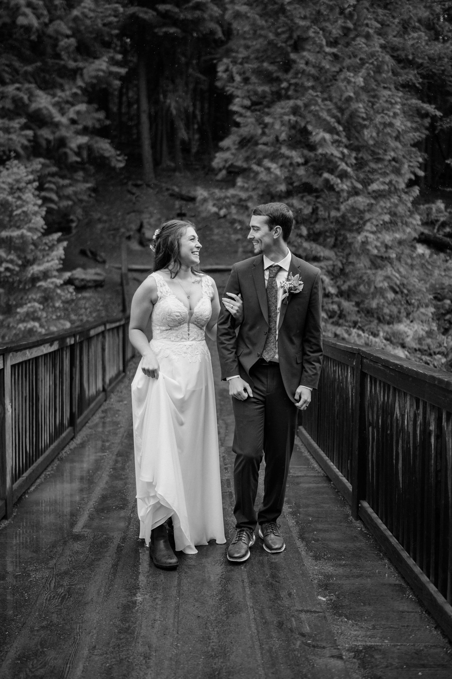 A couple in wedding attire shares playful moments on a wooden bridge surrounded by lush forest scenery.