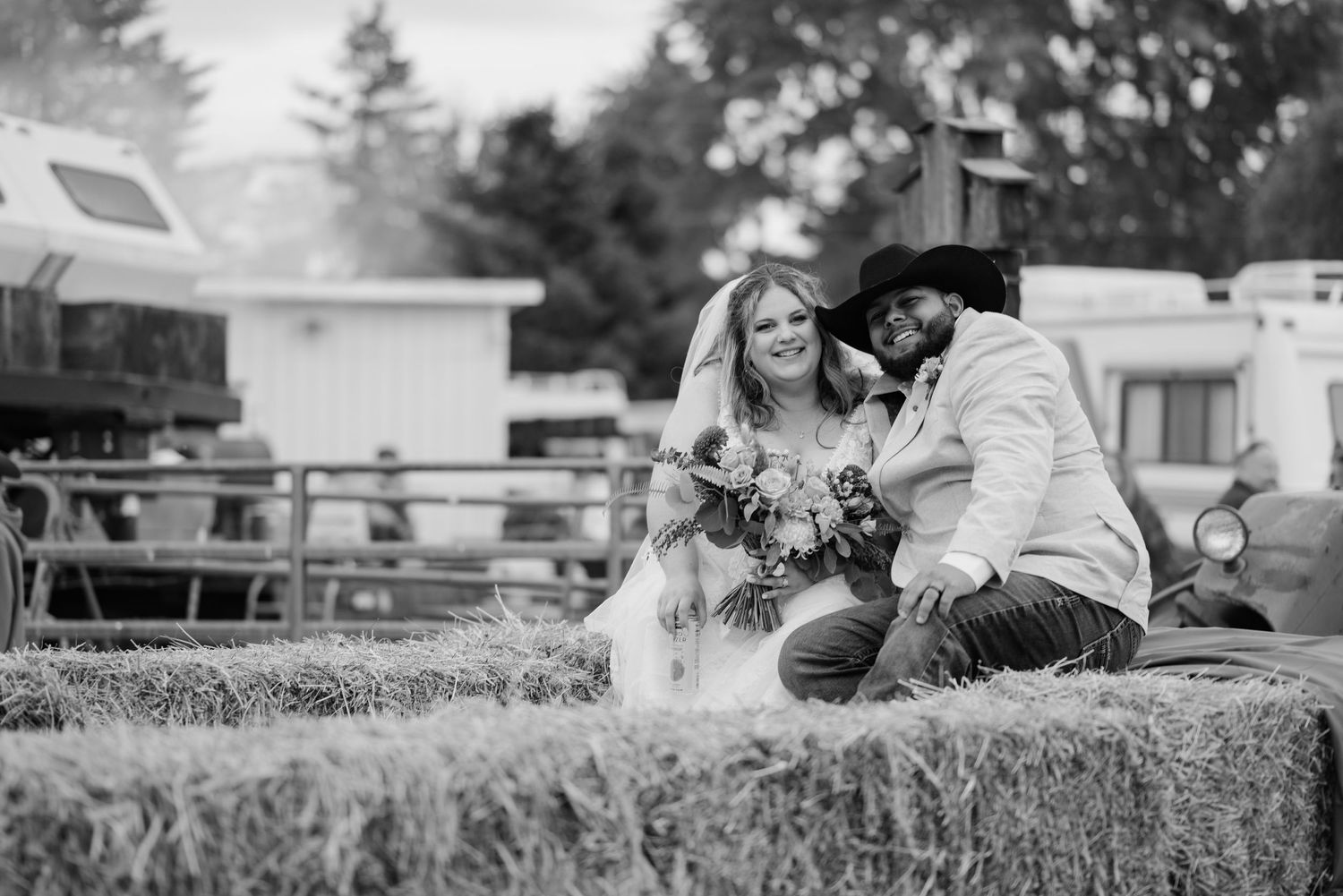 A black and white photograph shows a couple sitting together on hay bales outdoors at a rustic farm setting.