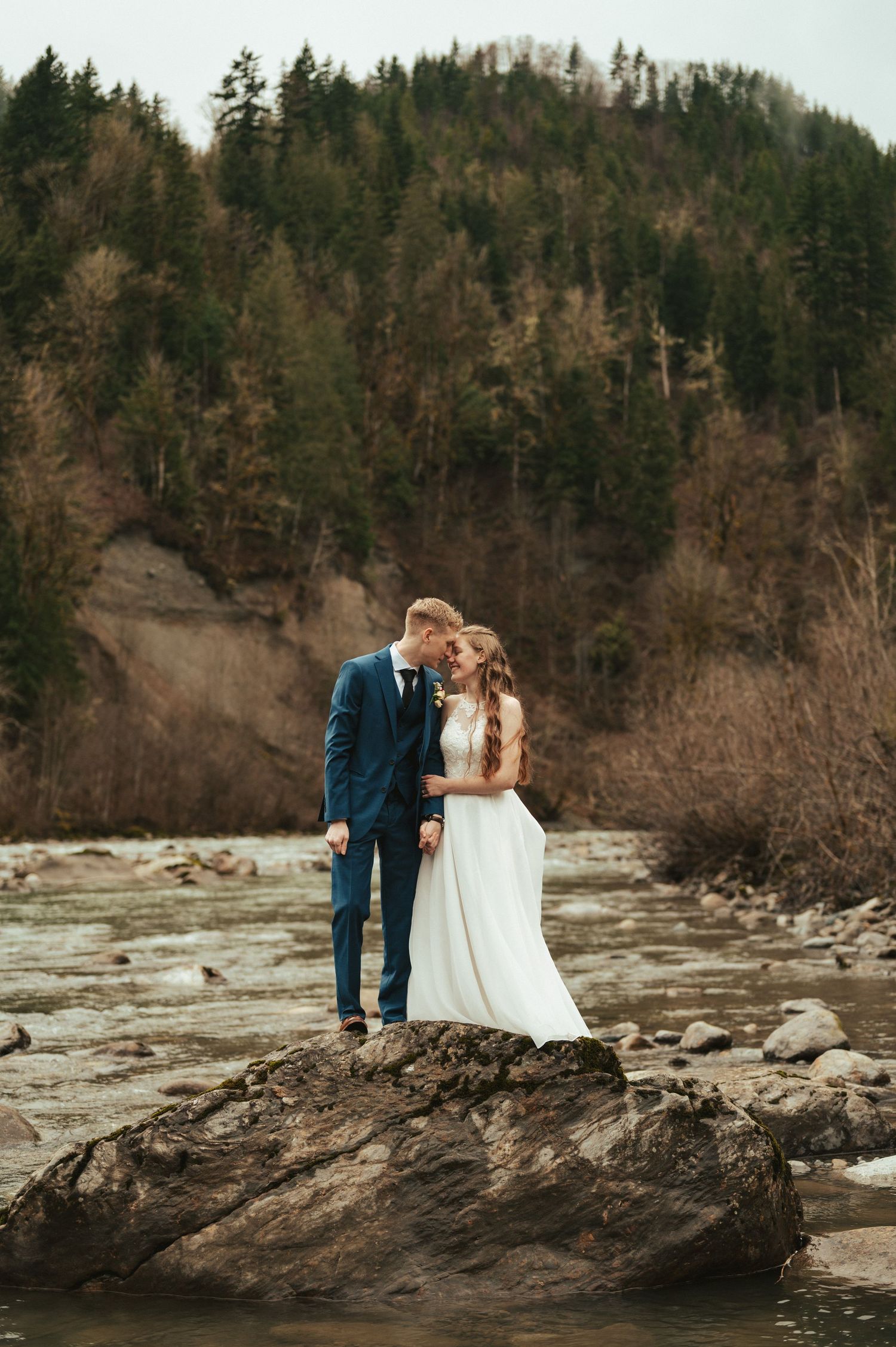 Couple shares intimate moment on rocky coastline with forested cliffs in background.