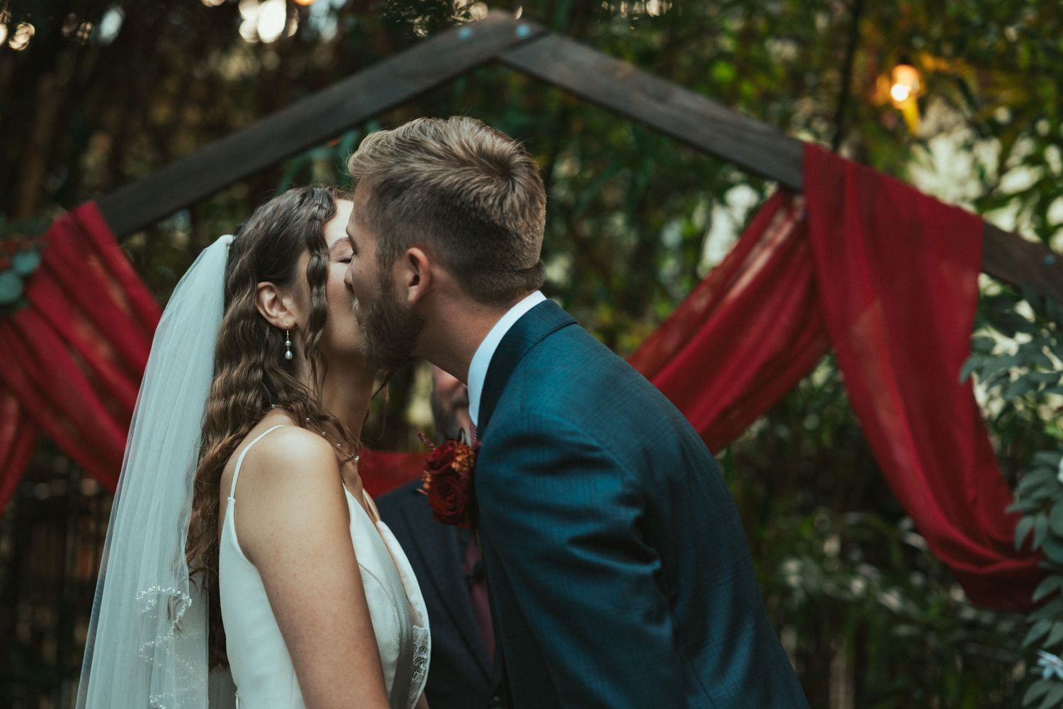 A couple shares a romantic kiss at their wedding ceremony with red fabric decor and greenery in the background.
