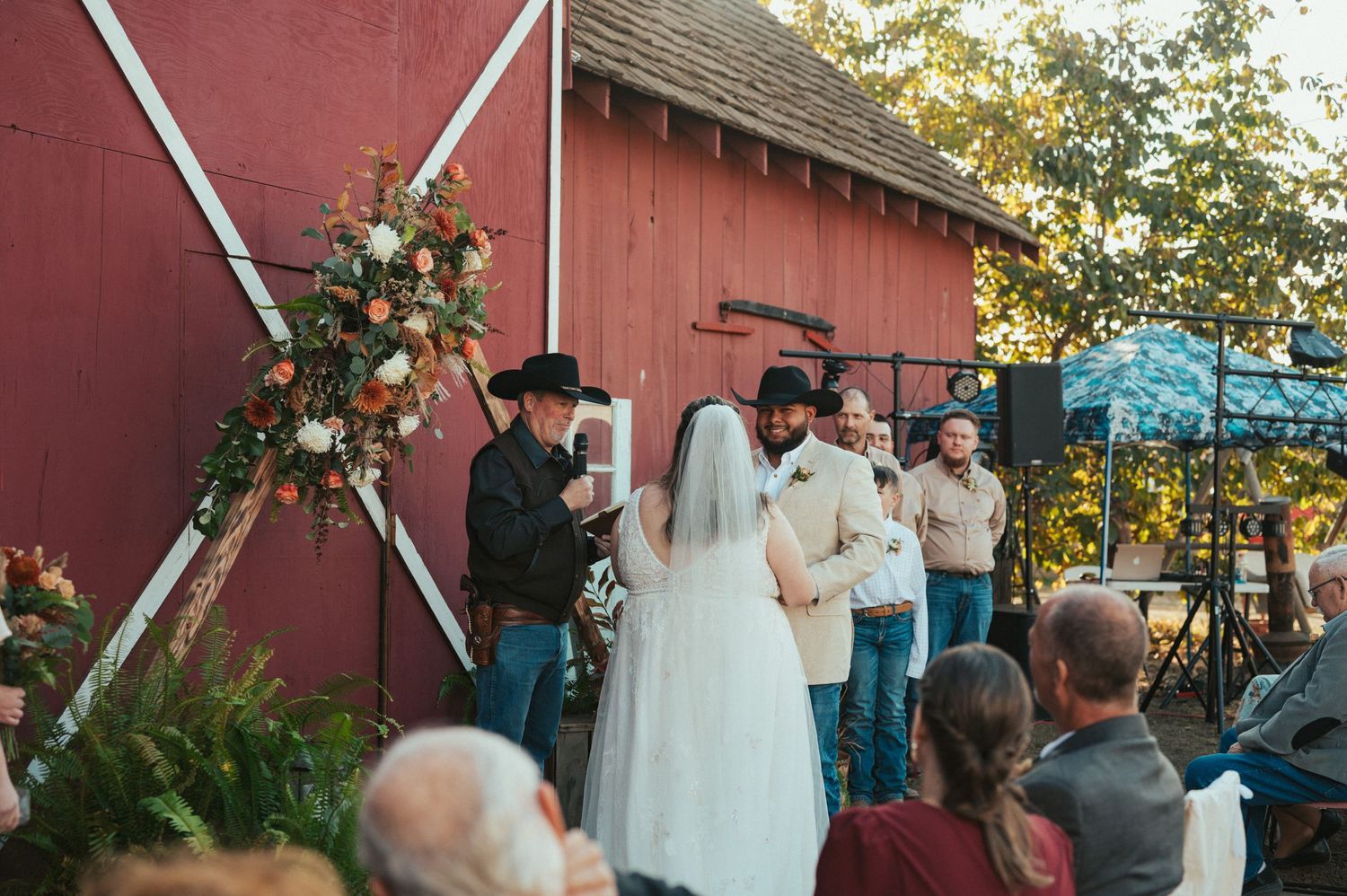 A wedding ceremony takes place in front of a rustic red barn with flower decorations on a sunny autumn day.