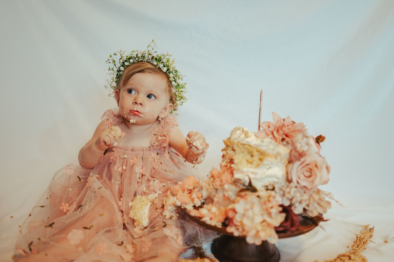 A baby wearing a flower crown and pink tulle dress reaches toward a decorated cake.