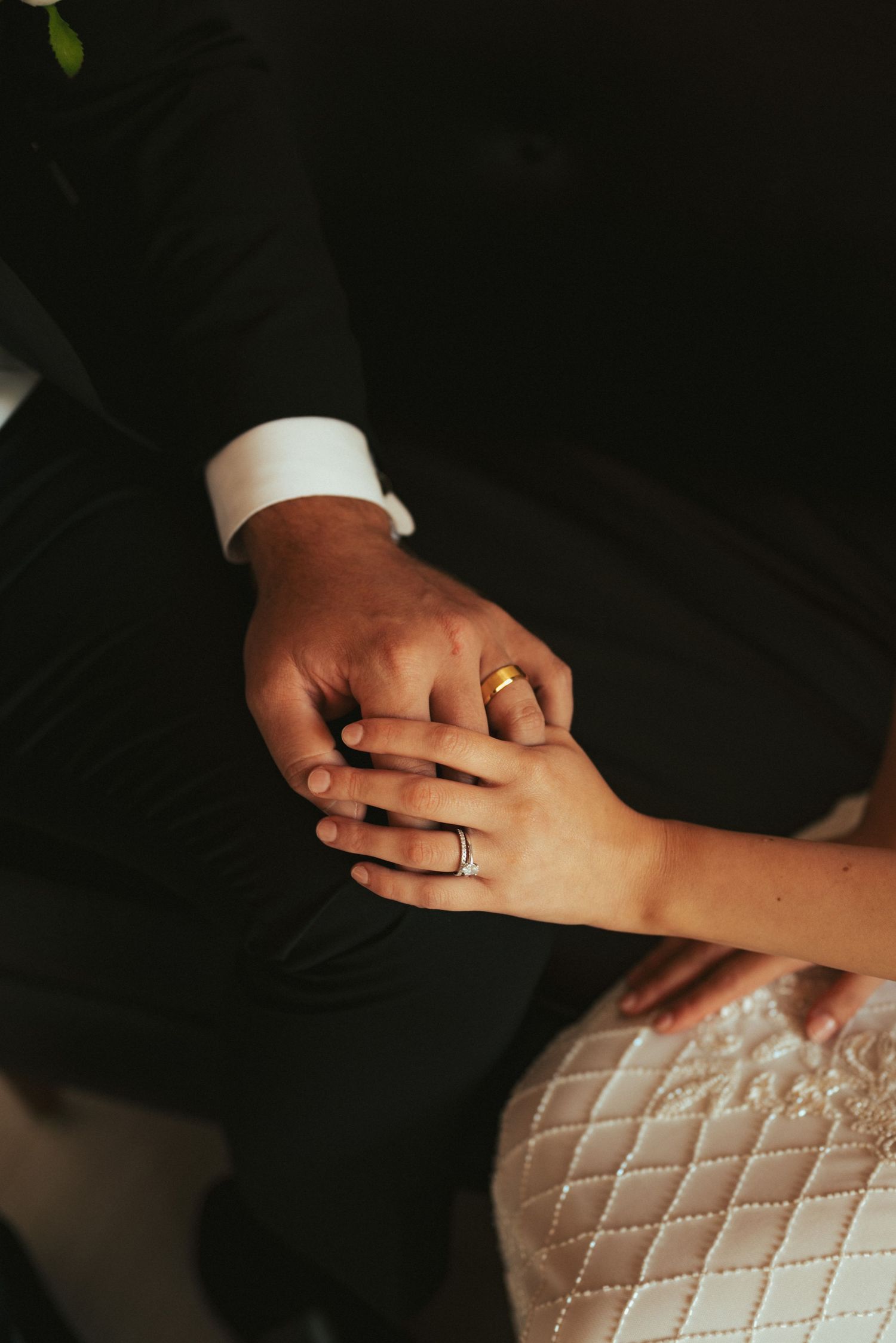 Close-up of newlyweds' hands with wedding rings against dark formal attire.