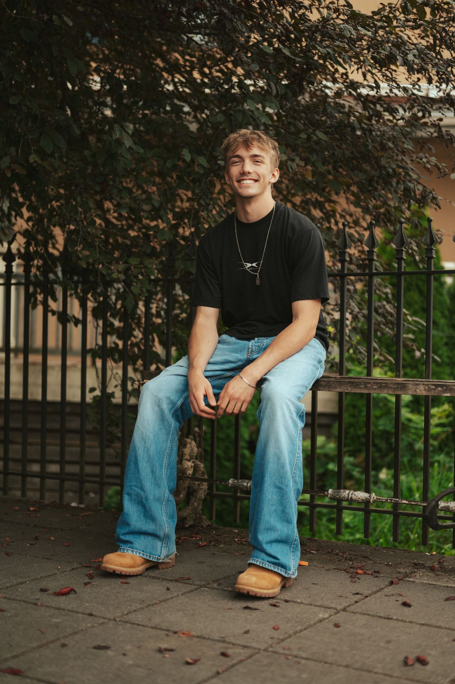 A casual portrait of someone in jeans and a black t-shirt sitting outdoors near autumn foliage.