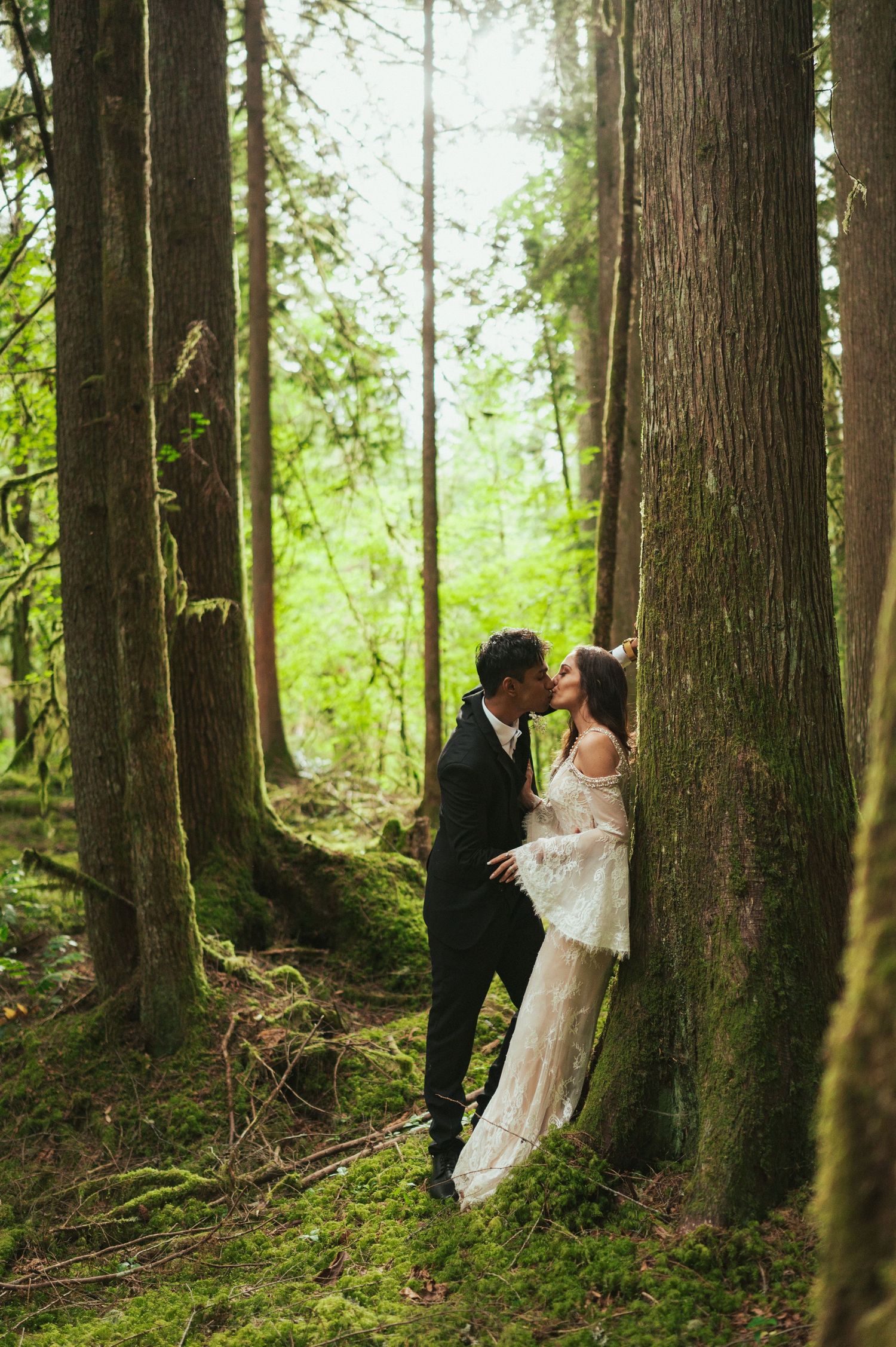 Wedding couple shares a romantic kiss among tall forest trees with sunlight streaming through.