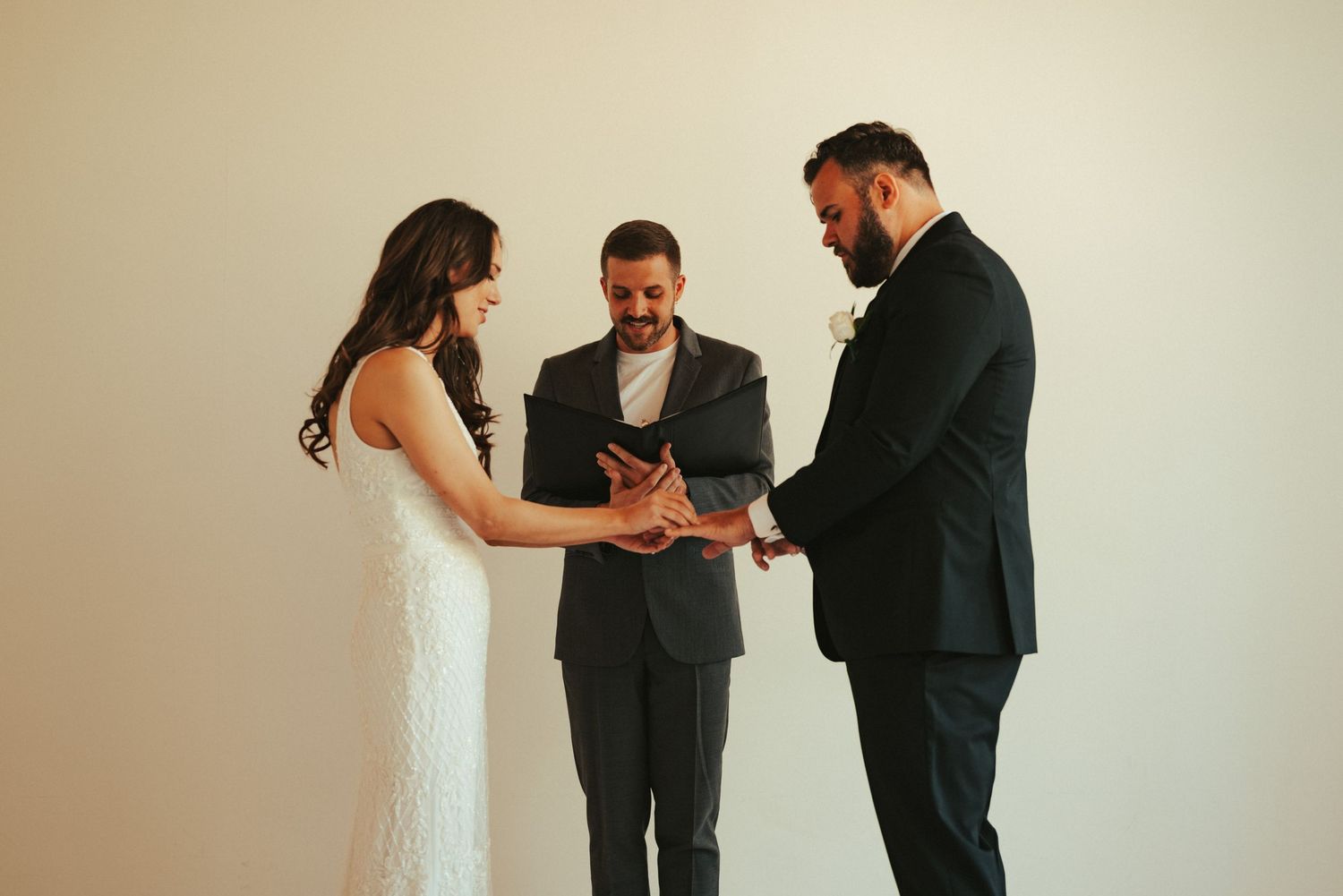 An intimate wedding ceremony with three people standing against a minimalist white wall.