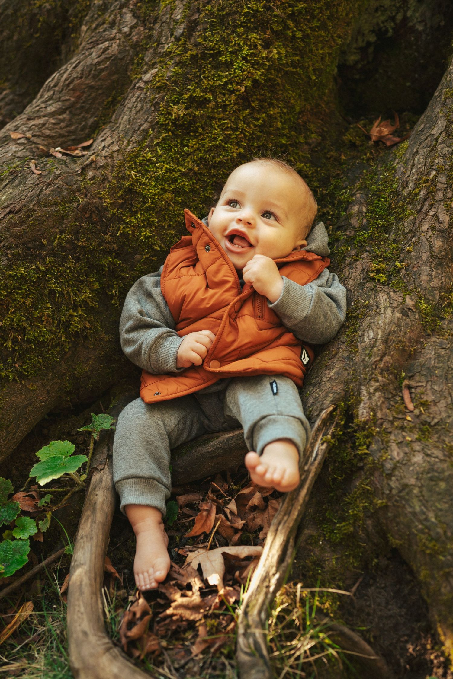 Baby resting against mossy tree trunk in orange and grey clothing during fall season.