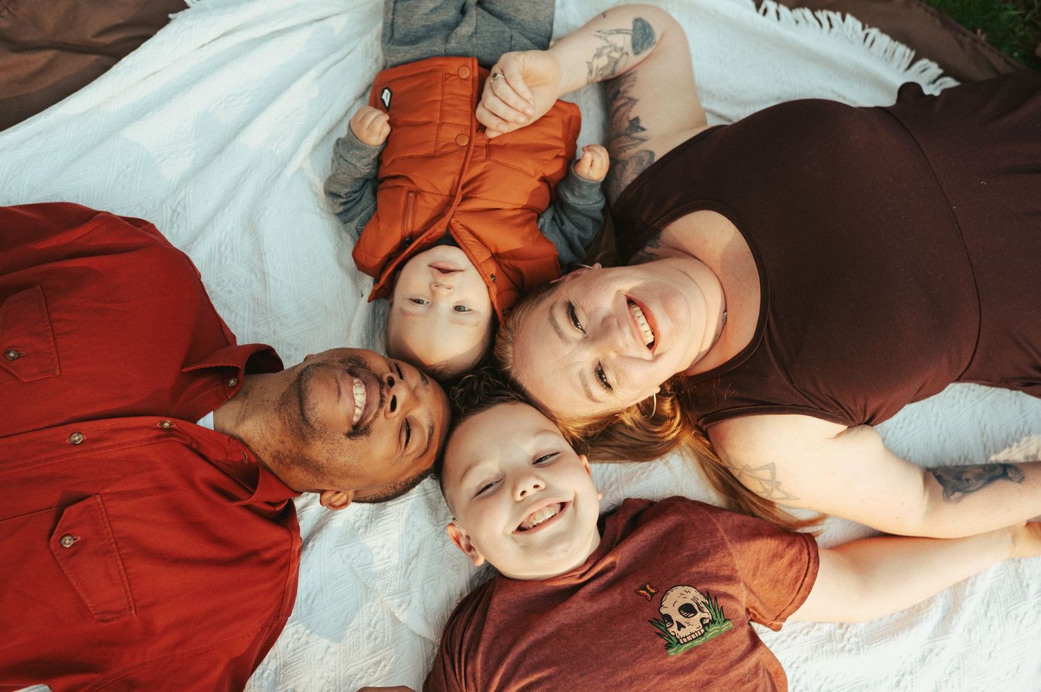 Four people lying in a circle with their heads together on a white blanket outdoors.