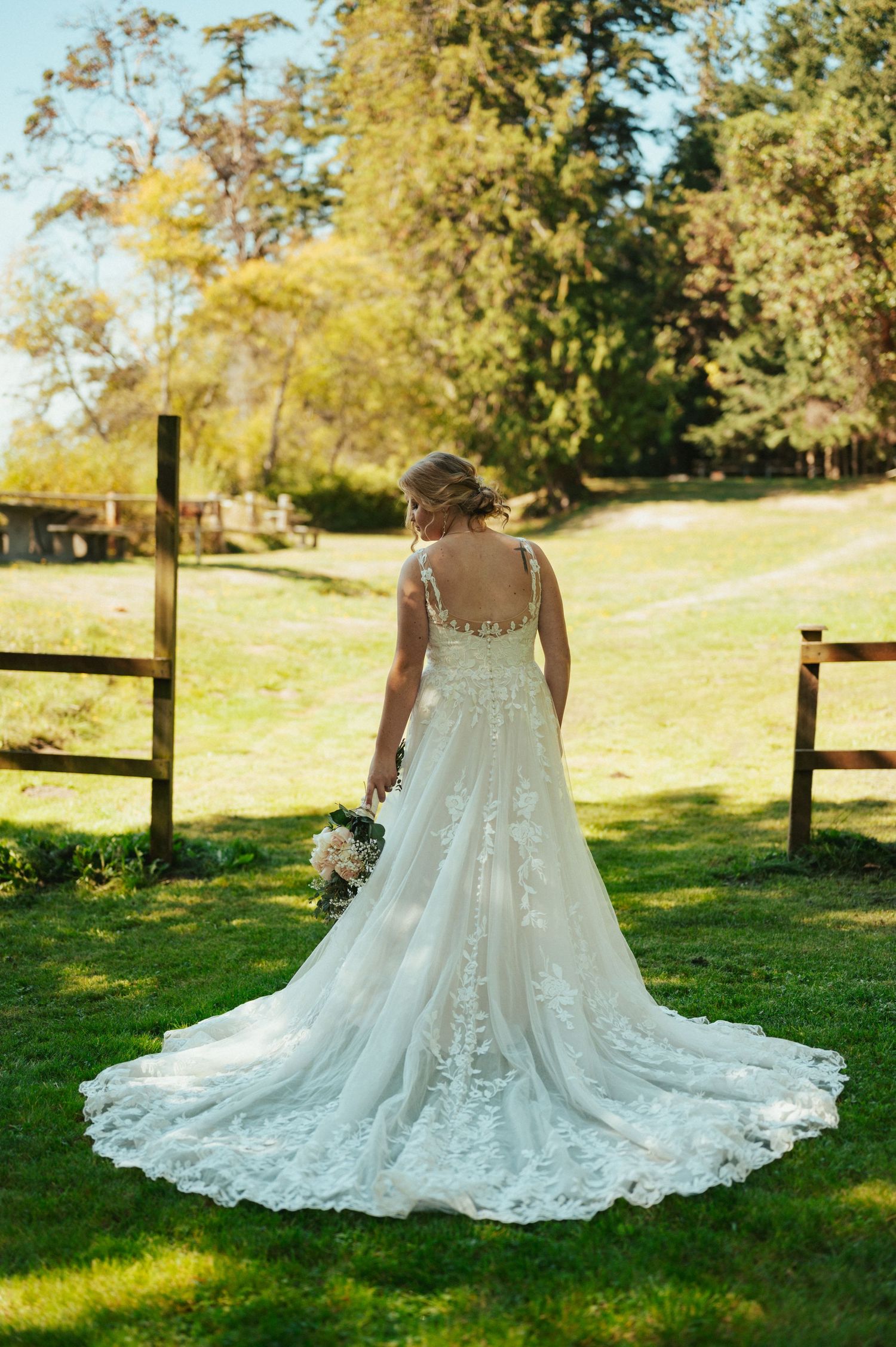 A detailed lace wedding dress with long train photographed from behind in a sunny garden setting.