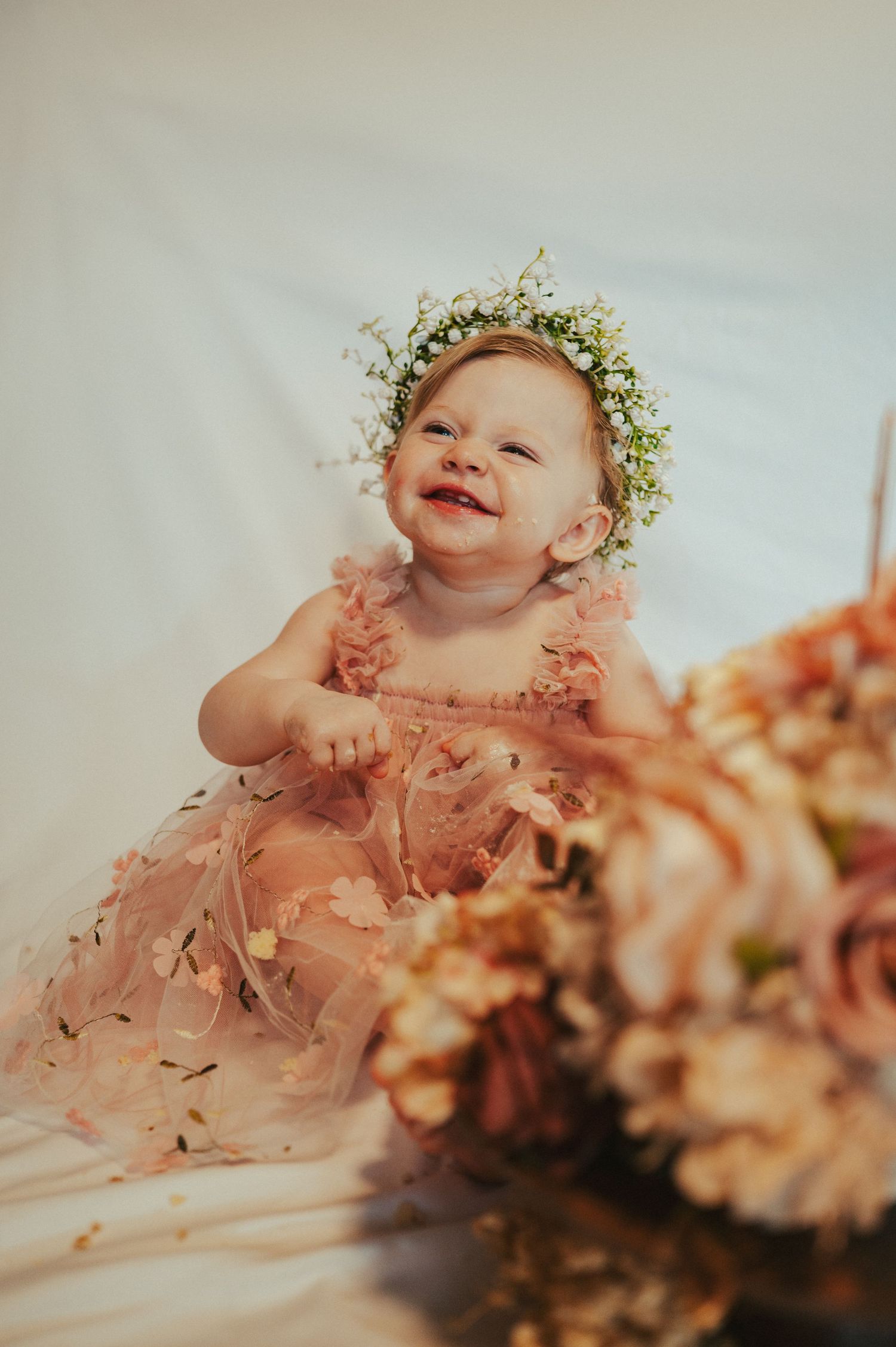 A baby wearing a floral crown and pink dress smiles during a cake smash photoshoot.