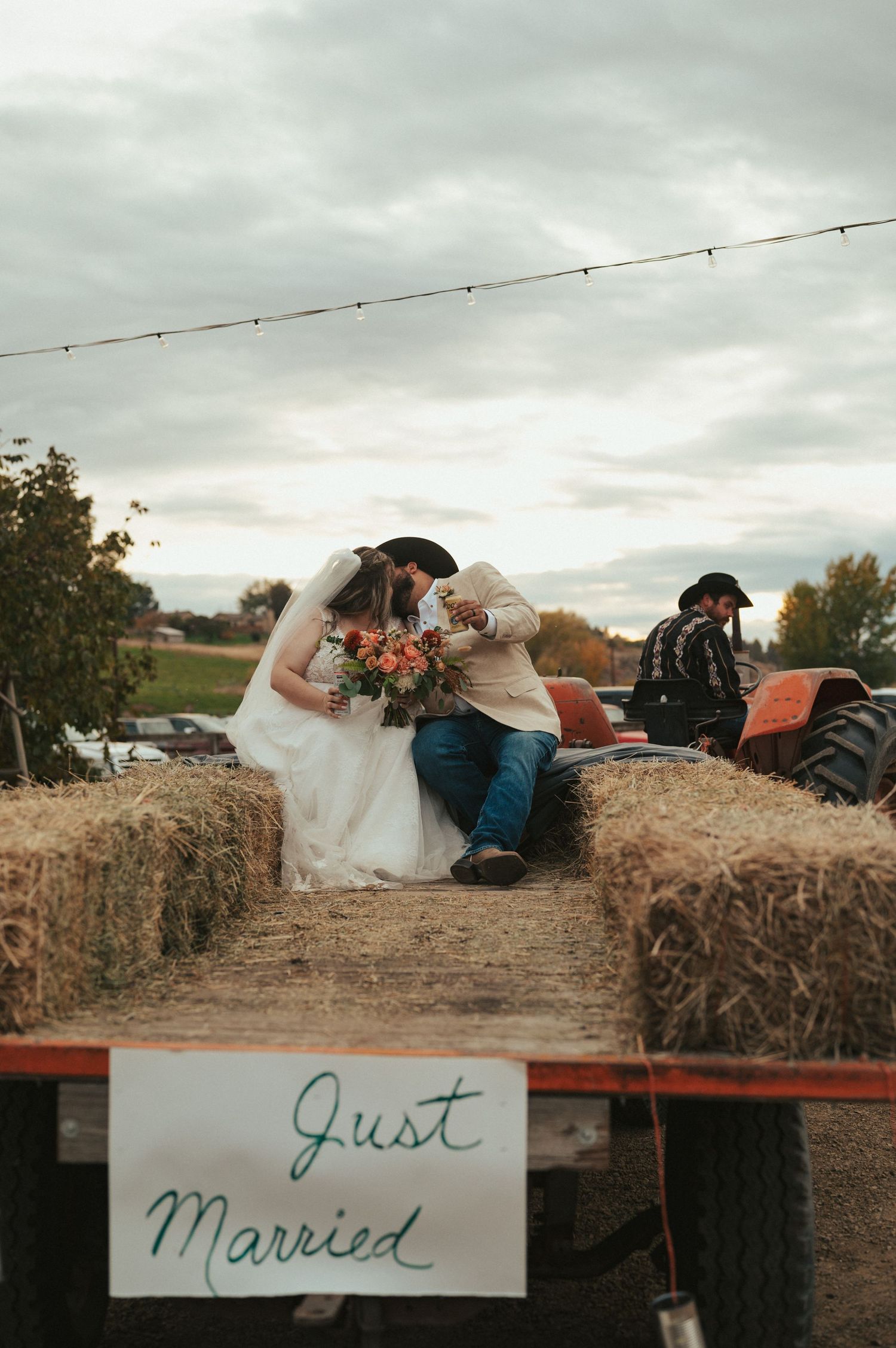 A couple sits on hay bales with a 'Just Married' sign during their rustic outdoor farm wedding.