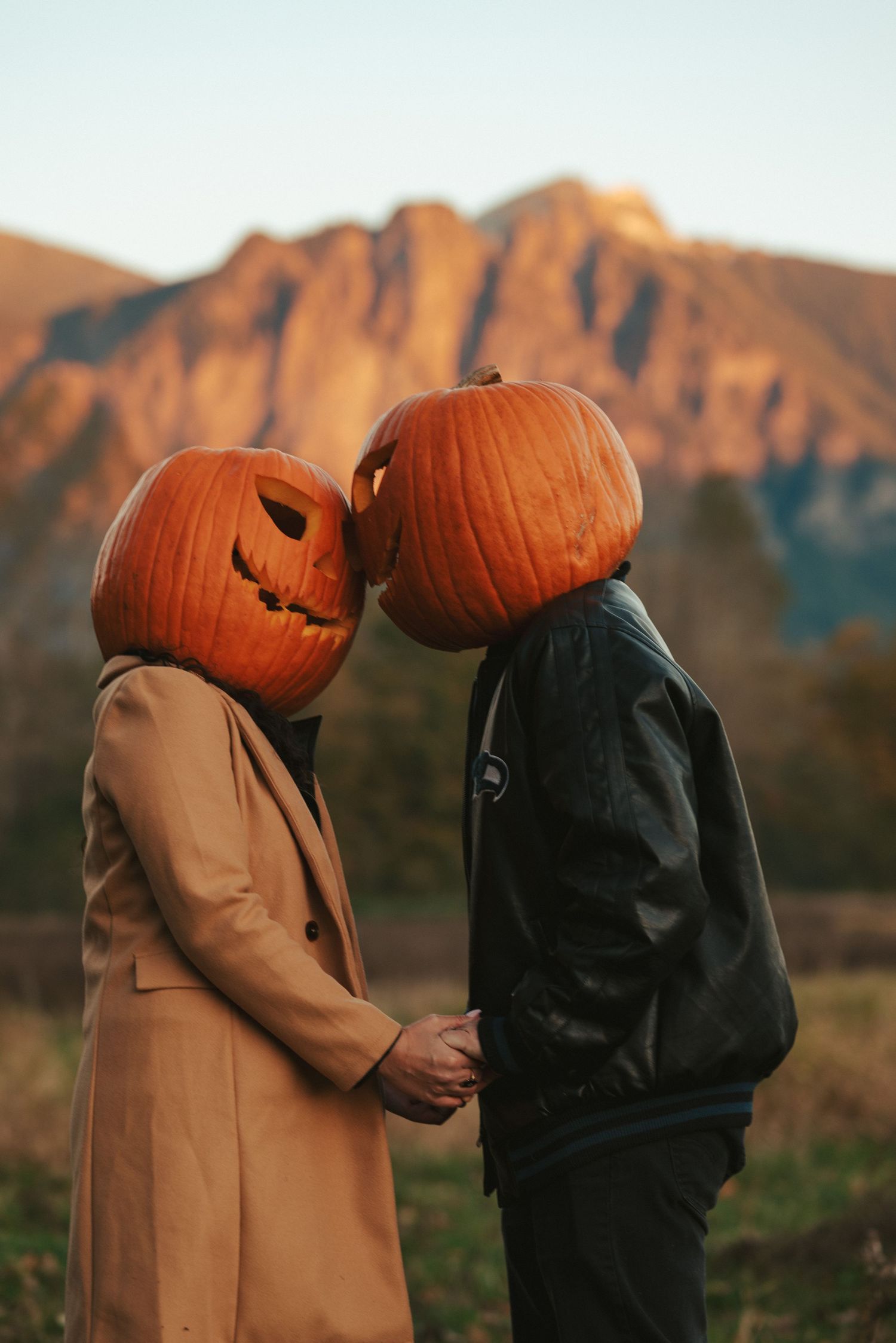 Two people wearing carved pumpkin heads and autumn coats hold hands against a mountain backdrop at sunset.