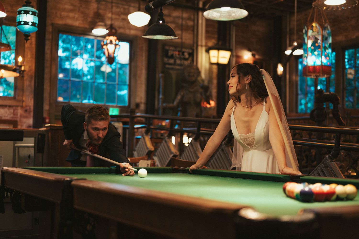 Someone in a white dress plays pool in a dimly lit bar with vintage lighting and blue-tinted windows.