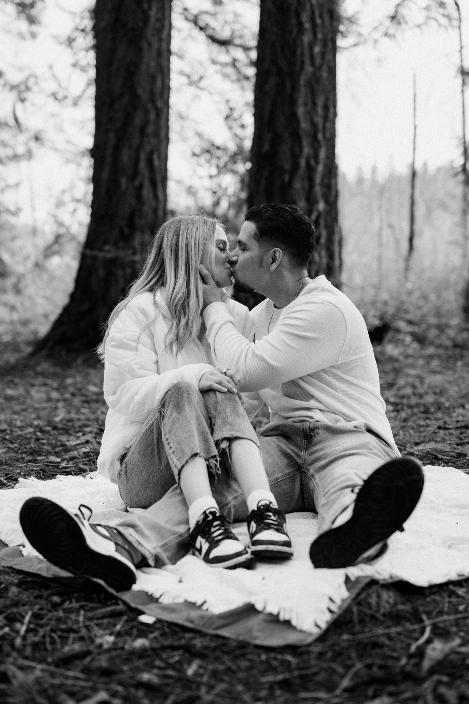 Black and white photo of couple sharing romantic moments on a blanket in a pine forest.