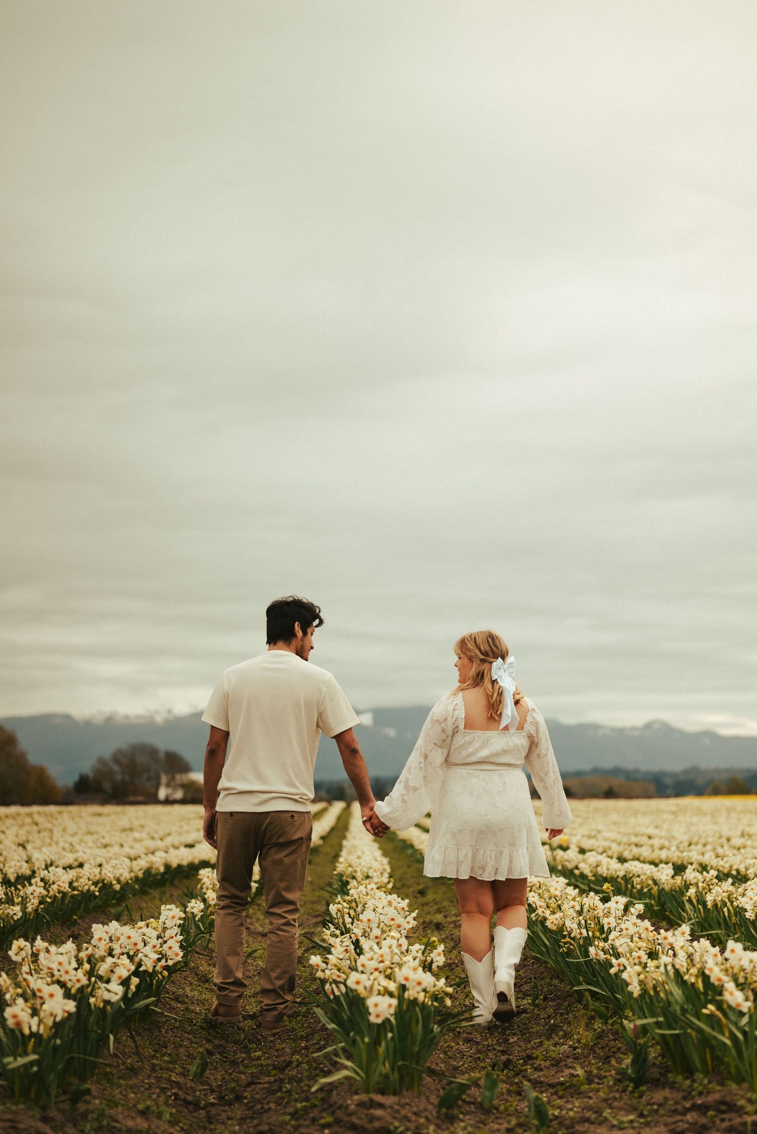A couple walks hand in hand through a field of blooming daffodils with mountains in the background.