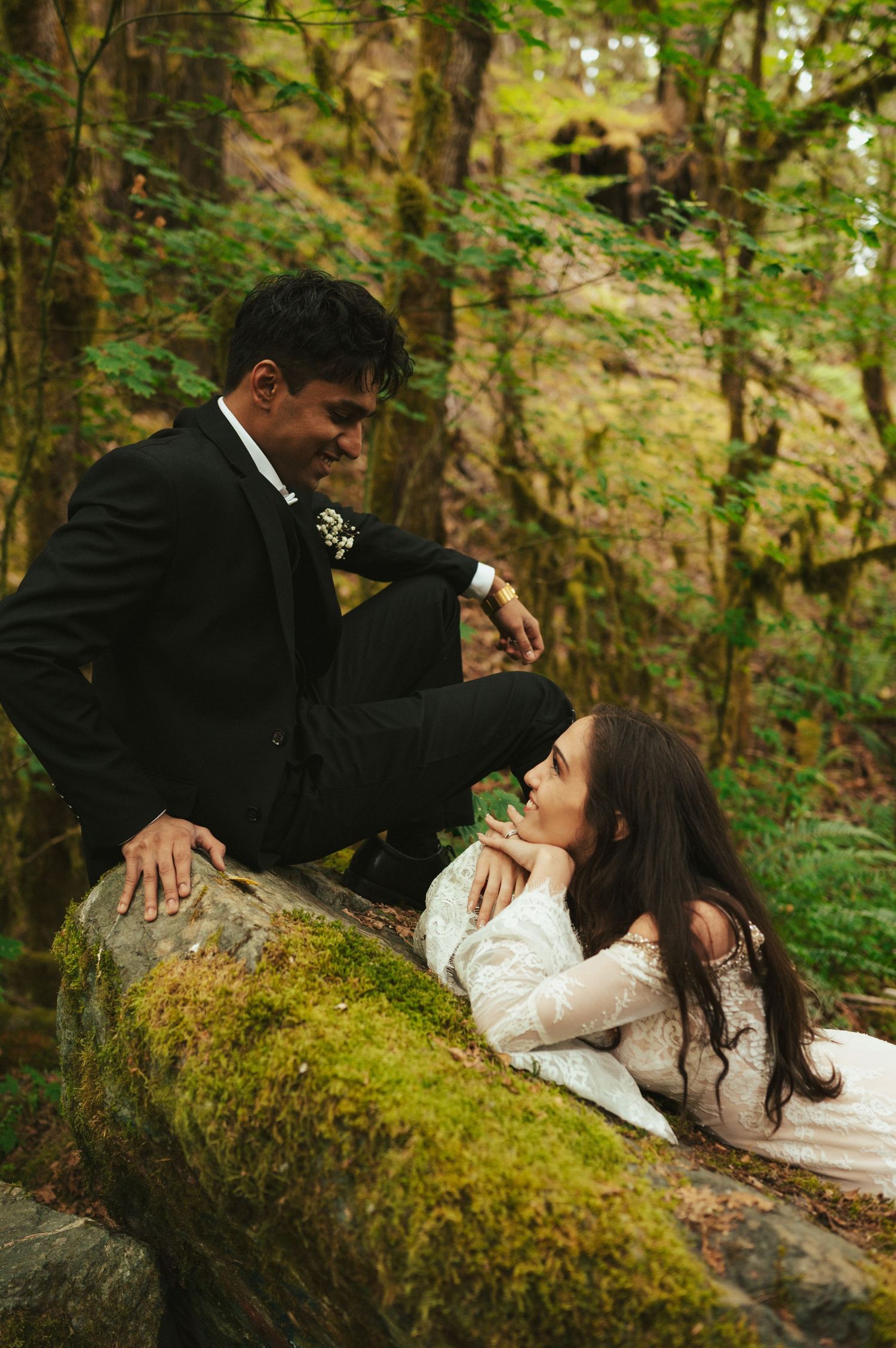 A couple in wedding attire poses intimately on a moss-covered log in a lush forest setting.