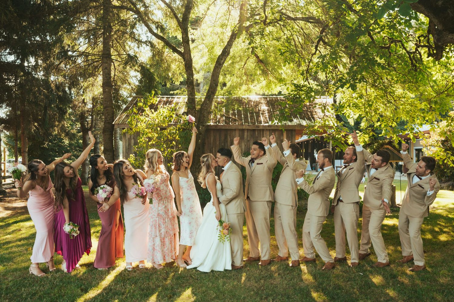 Wedding party poses outdoors in a garden with bridesmaids in pink dresses and groomsmen in beige suits.