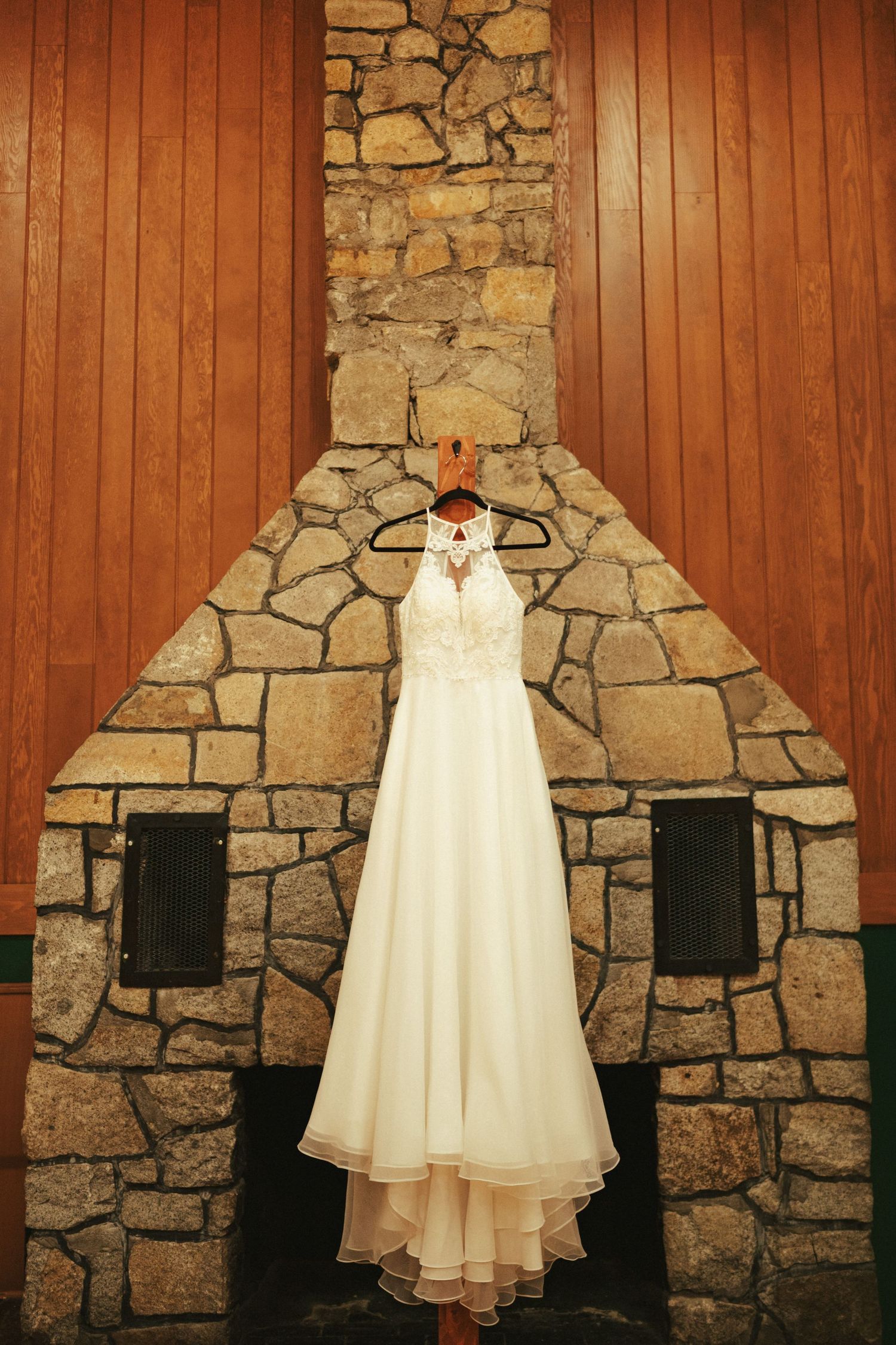 A white wedding dress hangs on a stone fireplace wall with wooden paneling on either side.