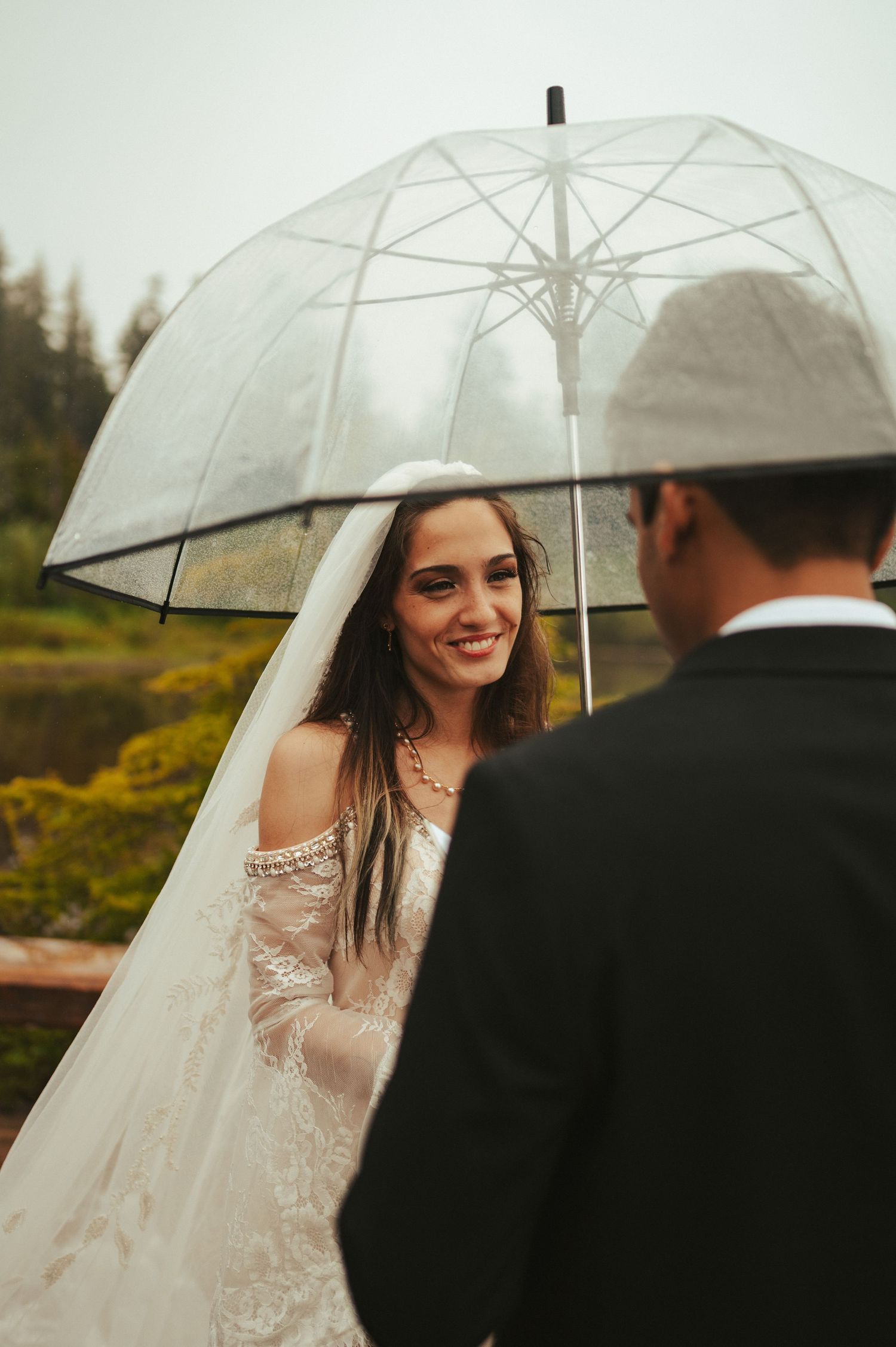 A bride under a clear umbrella smiles while wearing a lace wedding dress in an outdoor setting.