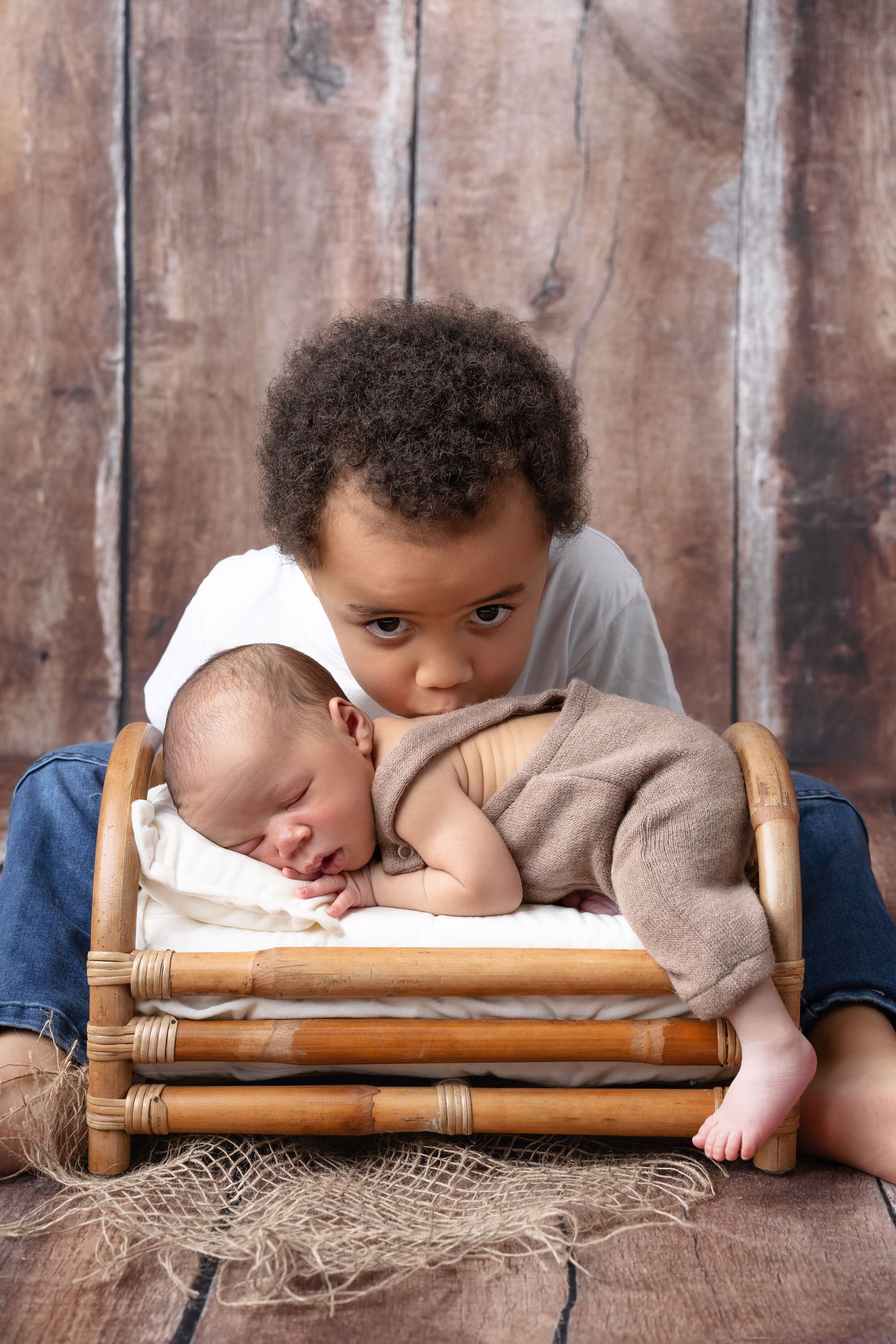 Young curly-haired toddler boy lovingly watching over his sleeping newborn sibling in a small wooden bed. séance naissance chavanoz lyon