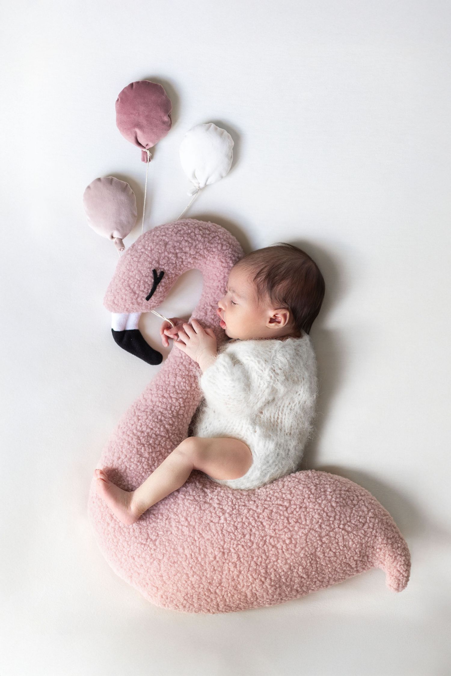 Sleeping newborn baby in white fuzzy outfit curled up next to a pink flamingo stuffed animal pillow.
séance naissance chavanoz lyon
