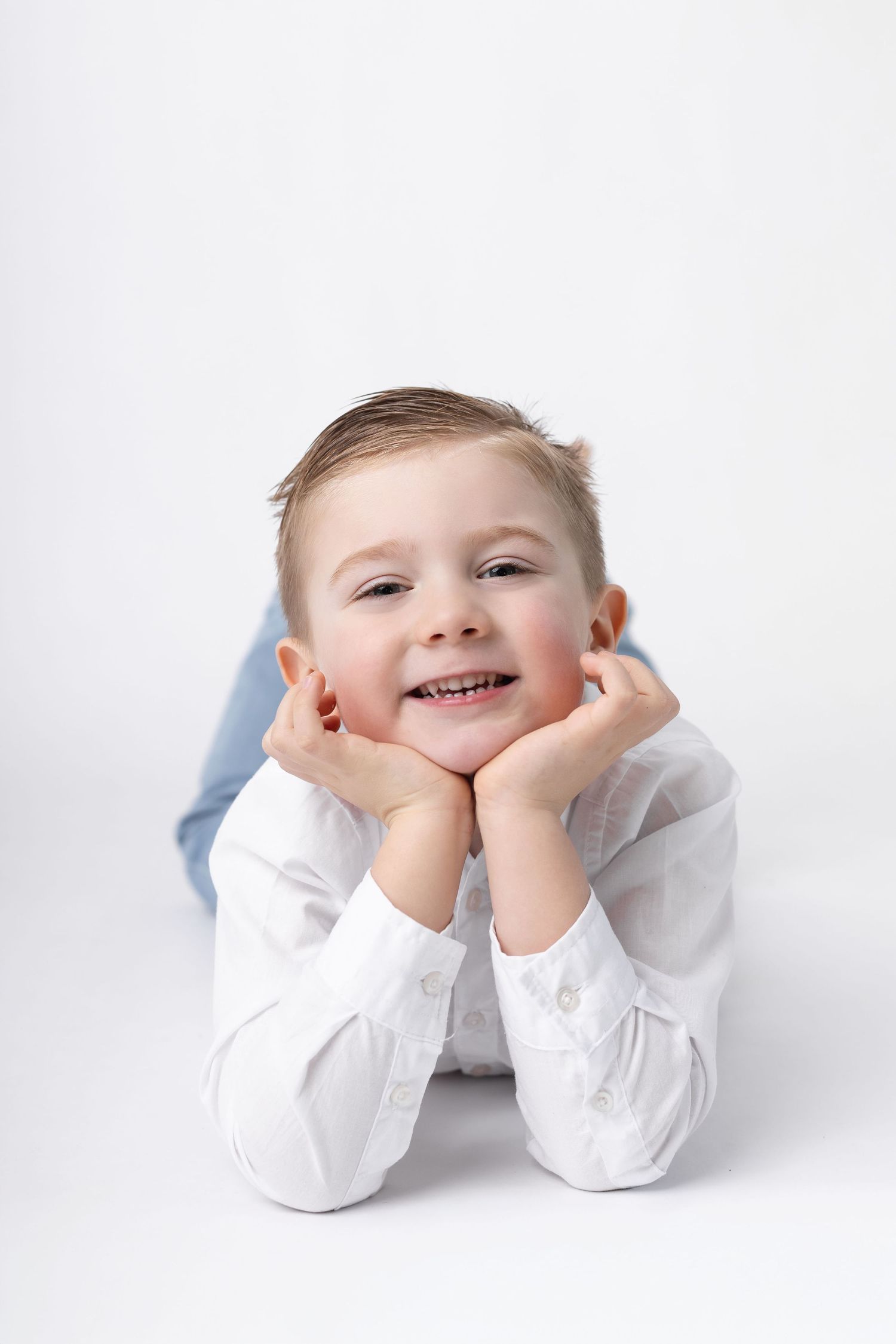 Smiling young boy in white shirt lies on floor with chin resting on hands against white background.