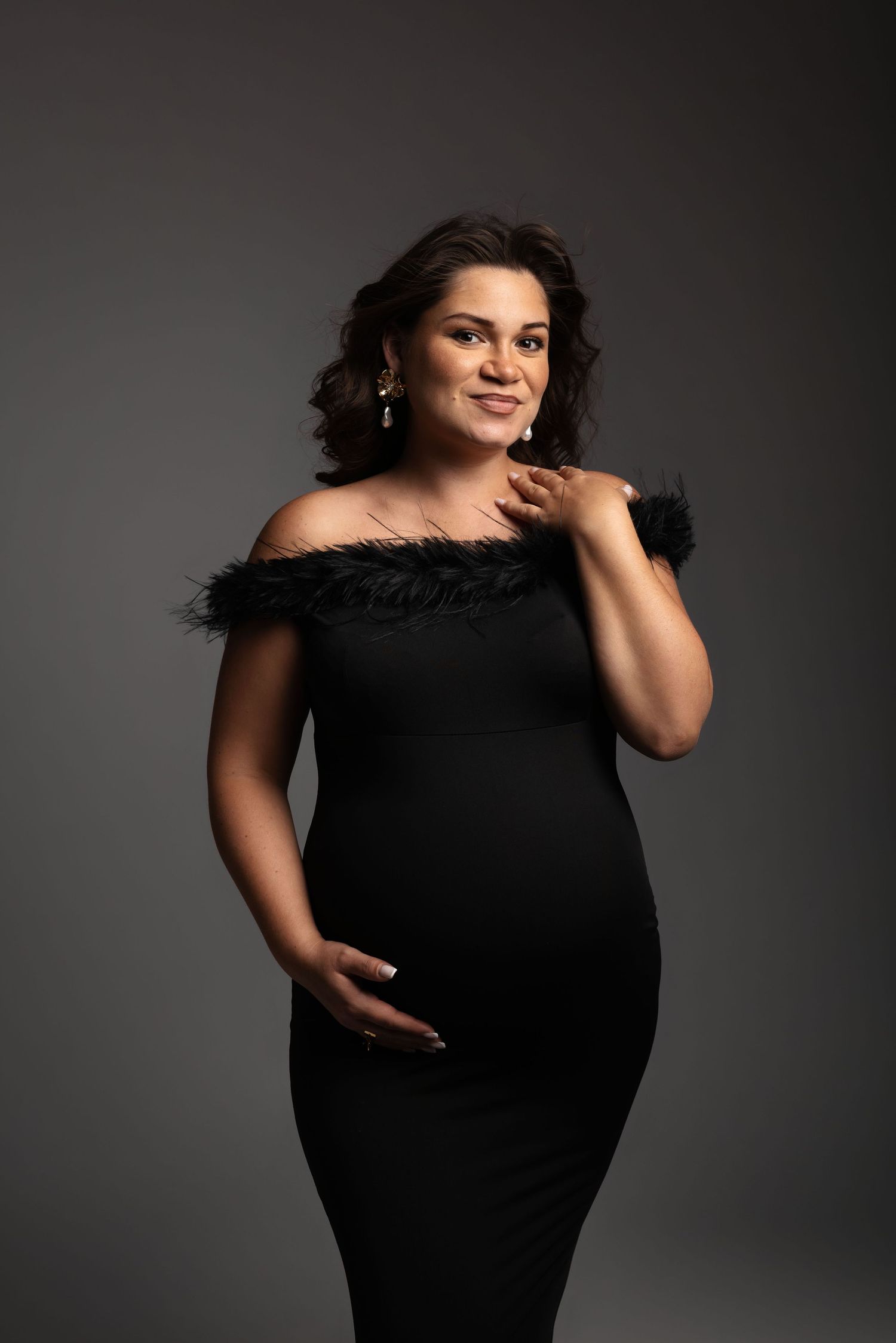 Pregnant woman in elegant black off-shoulder feathered dress posing gracefully against gray studio background.